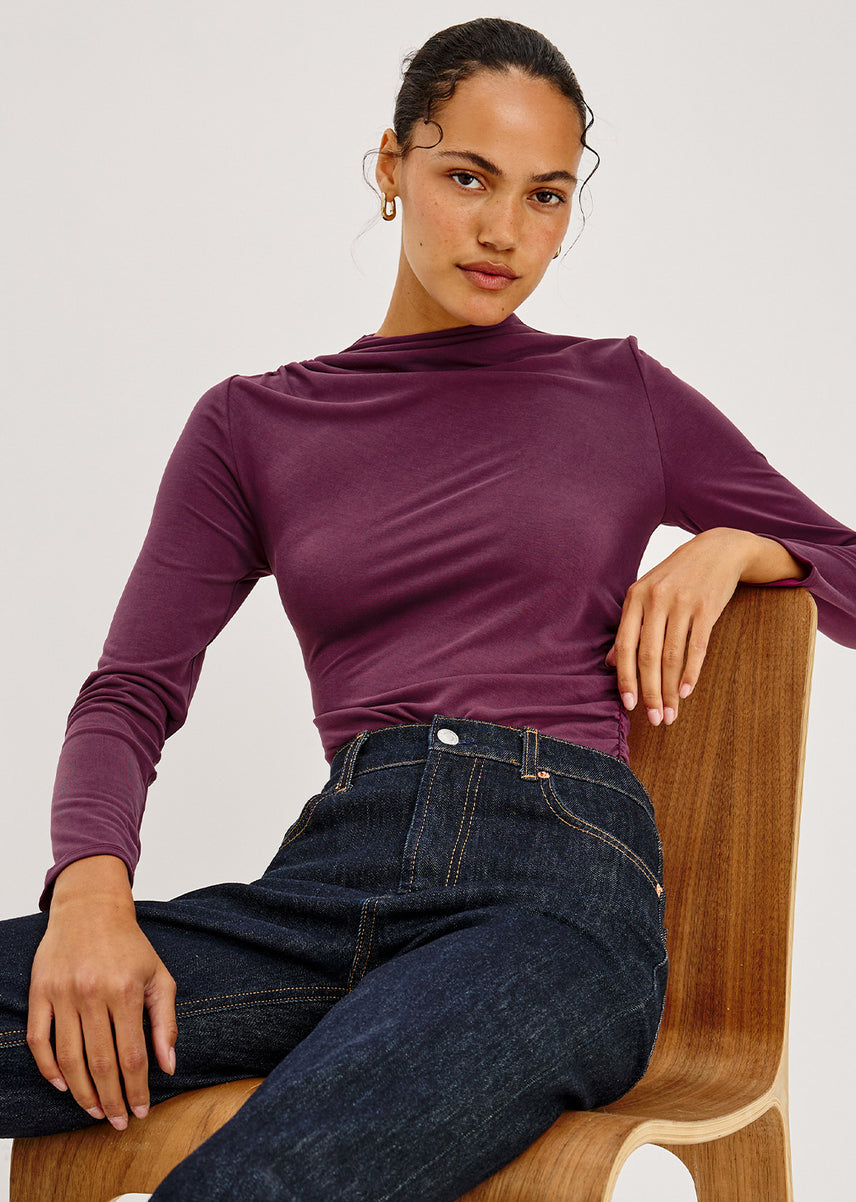 A close-up of a model in a plum draped, ruched mock-neck long-sleeve top and dark rinse denim, seated on a curved wooden chair, with small gold hoop earrings against a light gray background.