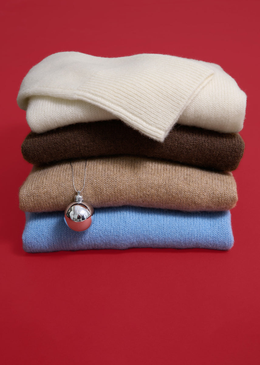 tack of four folded knit sweaters in cream, dark brown, camel, and light blue, with a silver ornament hanging from a thin chain against a red background.