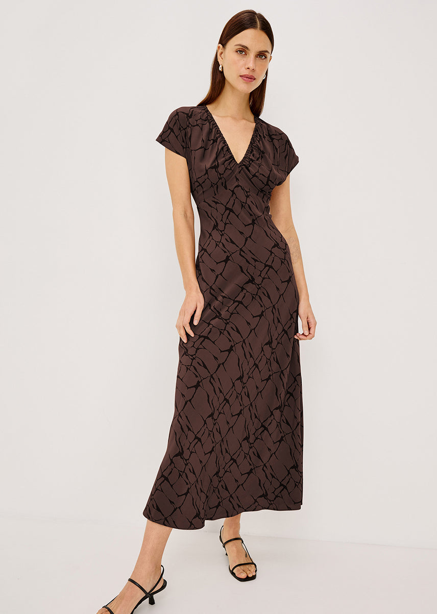 A woman modeling a dark brown midi wrap dress with a black abstract pattern, short flutter sleeves, and a V-neckline, set against a plain light gray studio background.