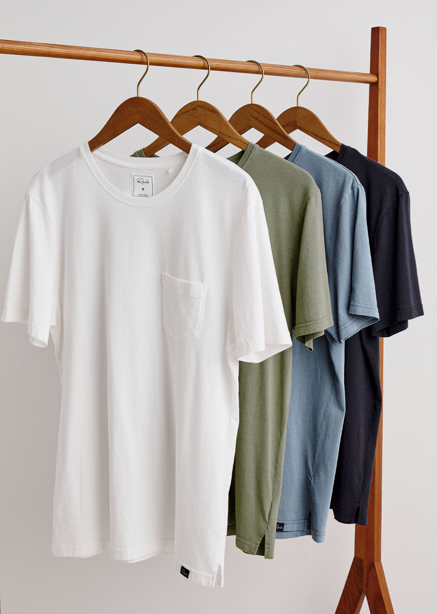 A set of short-sleeve crewneck pocket t-shirts hanging on wooden hangers from a wooden clothing rack. The shirts are shown in white, sage green, dusty blue, and navy blue.
