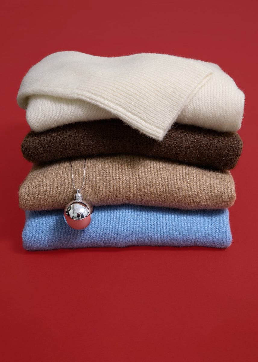 Stack of 4 colorful sweaters with an ornament hanging down placed against a red background. 
