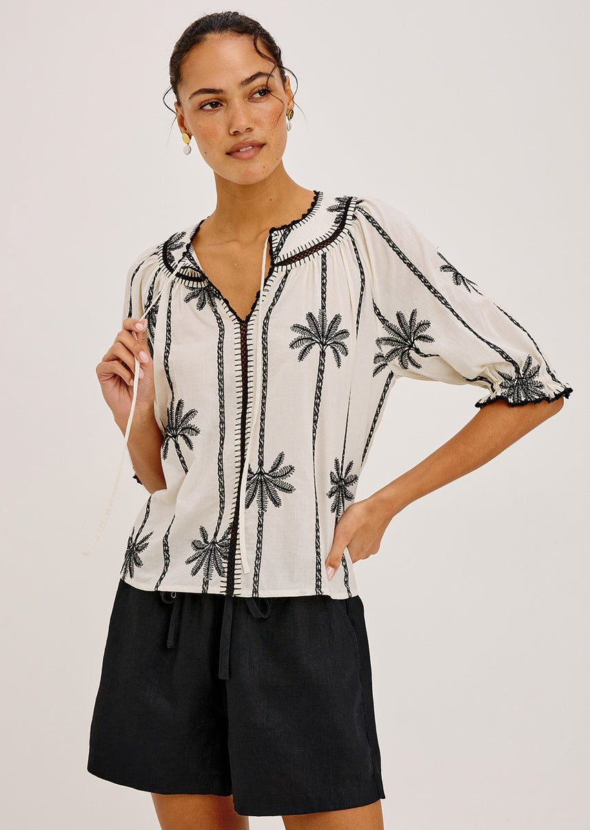 Woman wearing a white blouse with black floral embroidery and black shorts on a light gray background