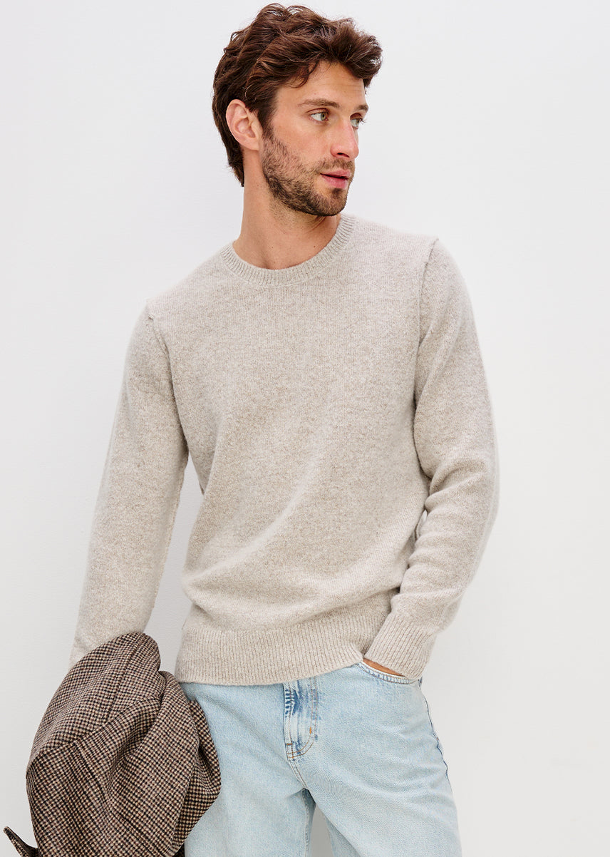 A man wearing a light beige crew-neck knit sweater and light blue jeans, holding a brown plaid jacket against a plain white background.