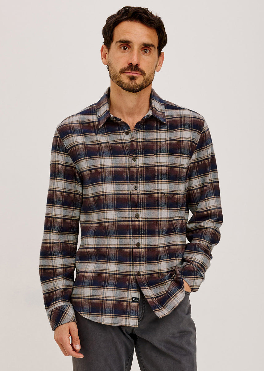 Man in a brown, navy, and gray plaid flannel shirt with gray pants, one hand in his pocket.