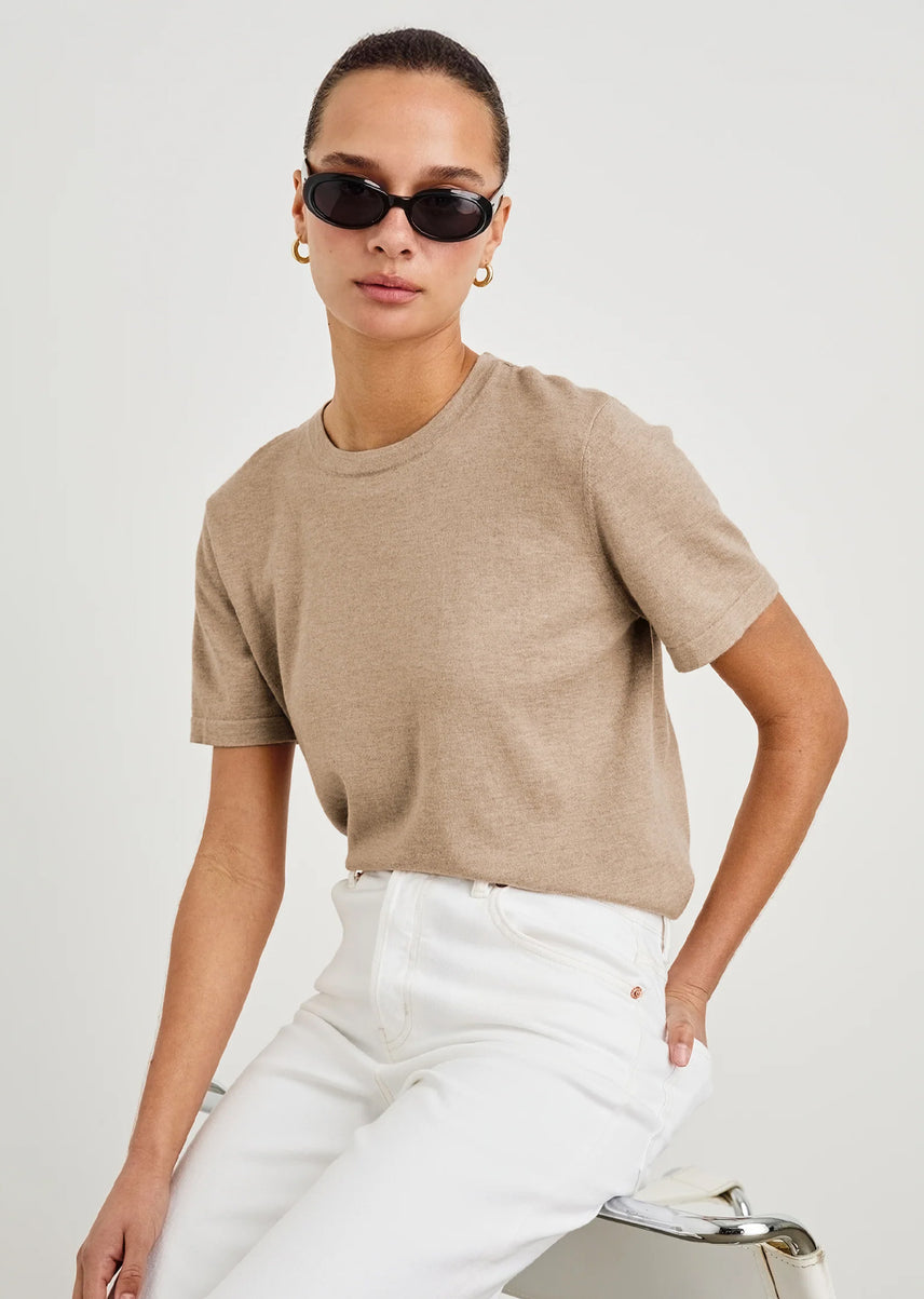 Woman wearing a beige t-shirt and white pants with sunglasses on a plain background