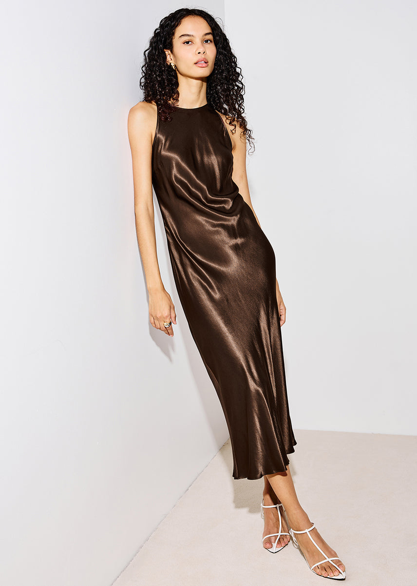 A woman modeling a sleeveless, high-neck satin midi dress in a rich dark brown color, paired with white strappy high-heeled sandals, set against a minimalist off-white studio background.