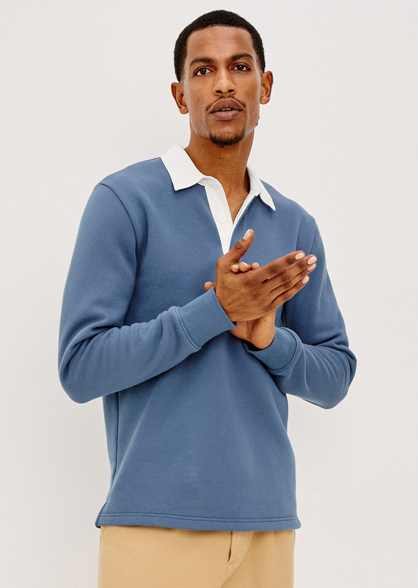 A man wearing a dusty blue long-sleeve polo shirt with a white collar, paired with beige trousers, standing against a light gray studio background.