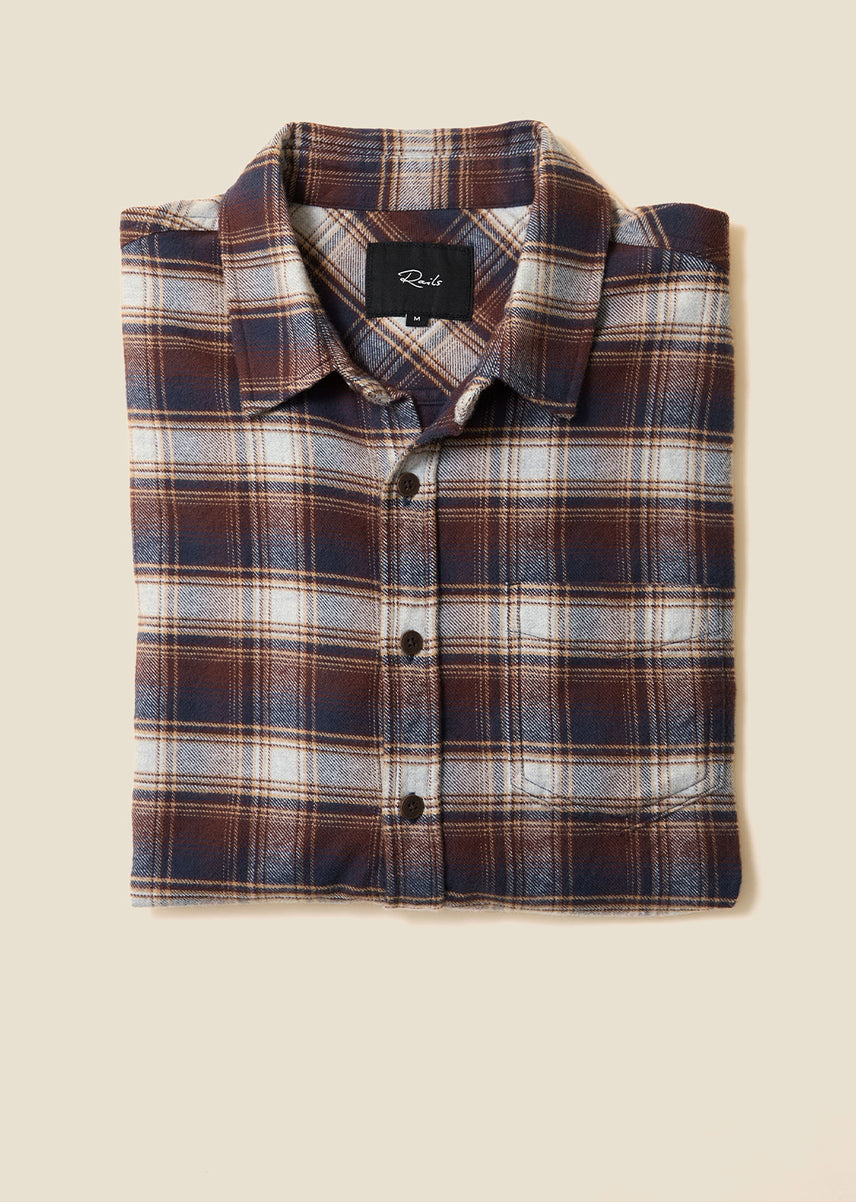 Folded flannel button-up shirt in brown, navy, and cream plaid with a chest pocket and a Rails label at the collar, on a beige background.