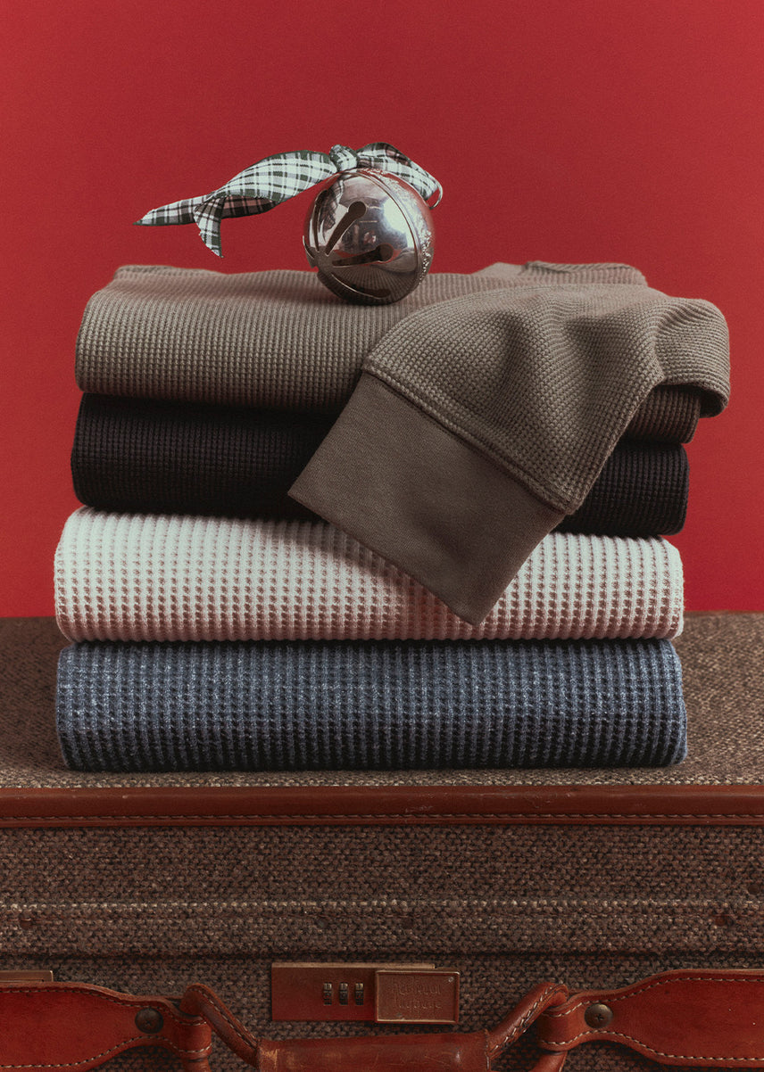 Stack of four waffle-knit thermal tops in taupe, black, pale pink, and blue, topped with a silver sleigh bell tied with a plaid ribbon, resting on a vintage tweed suitcase against a red wall.