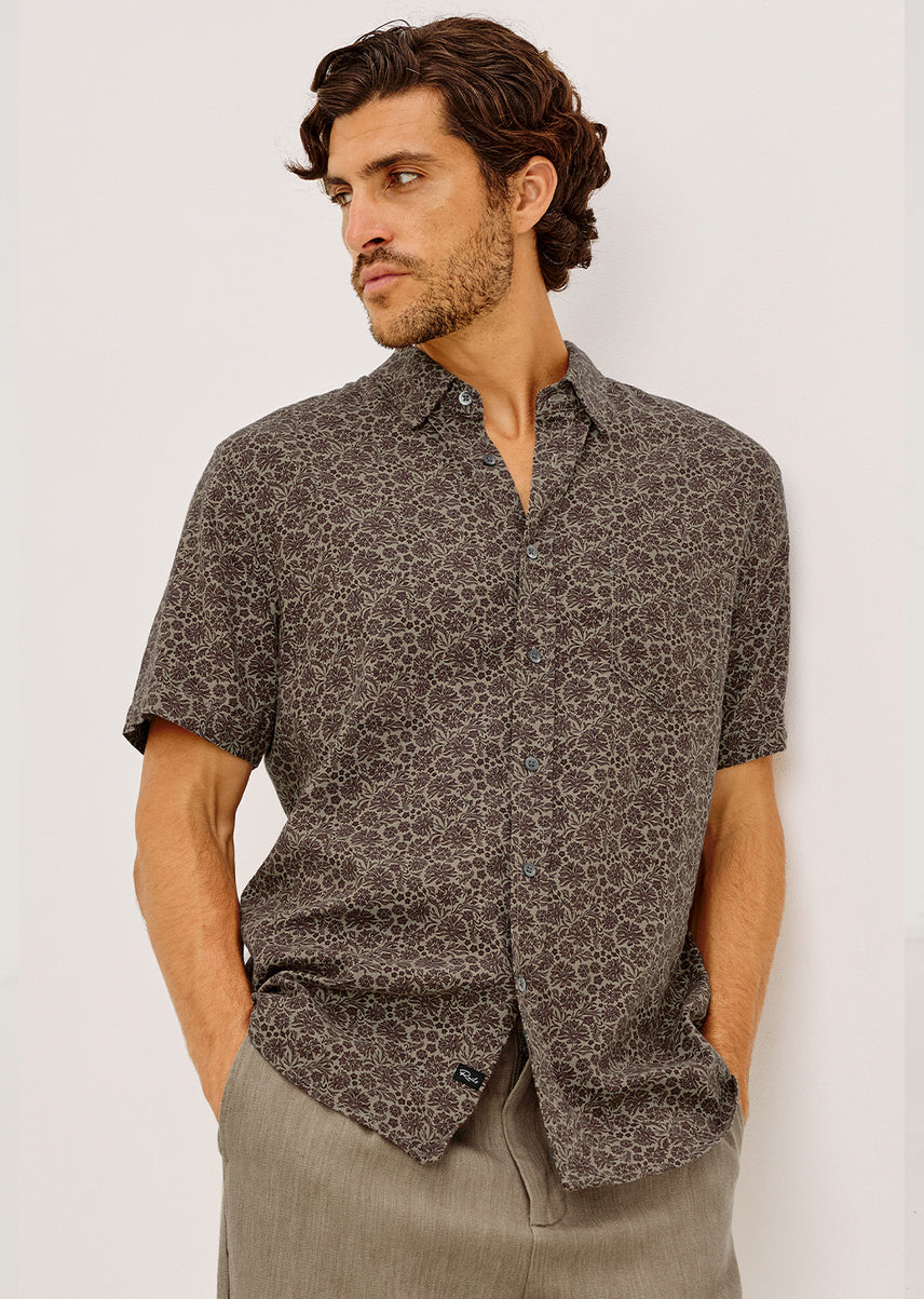 A man wearing a short-sleeve button-up shirt with a dark brown and gray abstract pattern, paired with light gray trousers, standing against a plain light gray background.