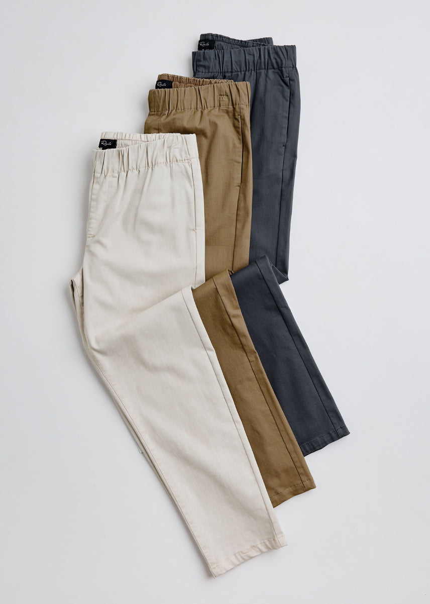 Four pairs of men's elastic-waist trousers in cream, tan, brown, and charcoal gray, arranged in a fan shape on a light gray background.