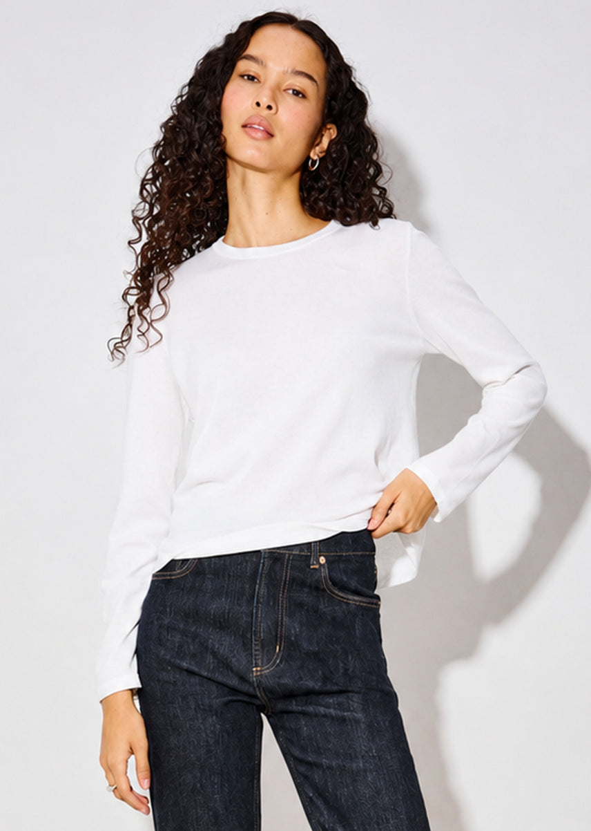 A woman with long curly dark hair wearing a white long-sleeve crewneck top and dark blue jeans, standing against a light gray studio background.
