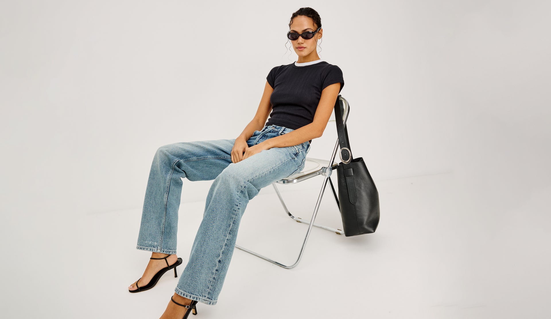 Woman sitting on a chair wearing a black top, blue jeans, and sunglasses with a black handbag beside her on a white background.