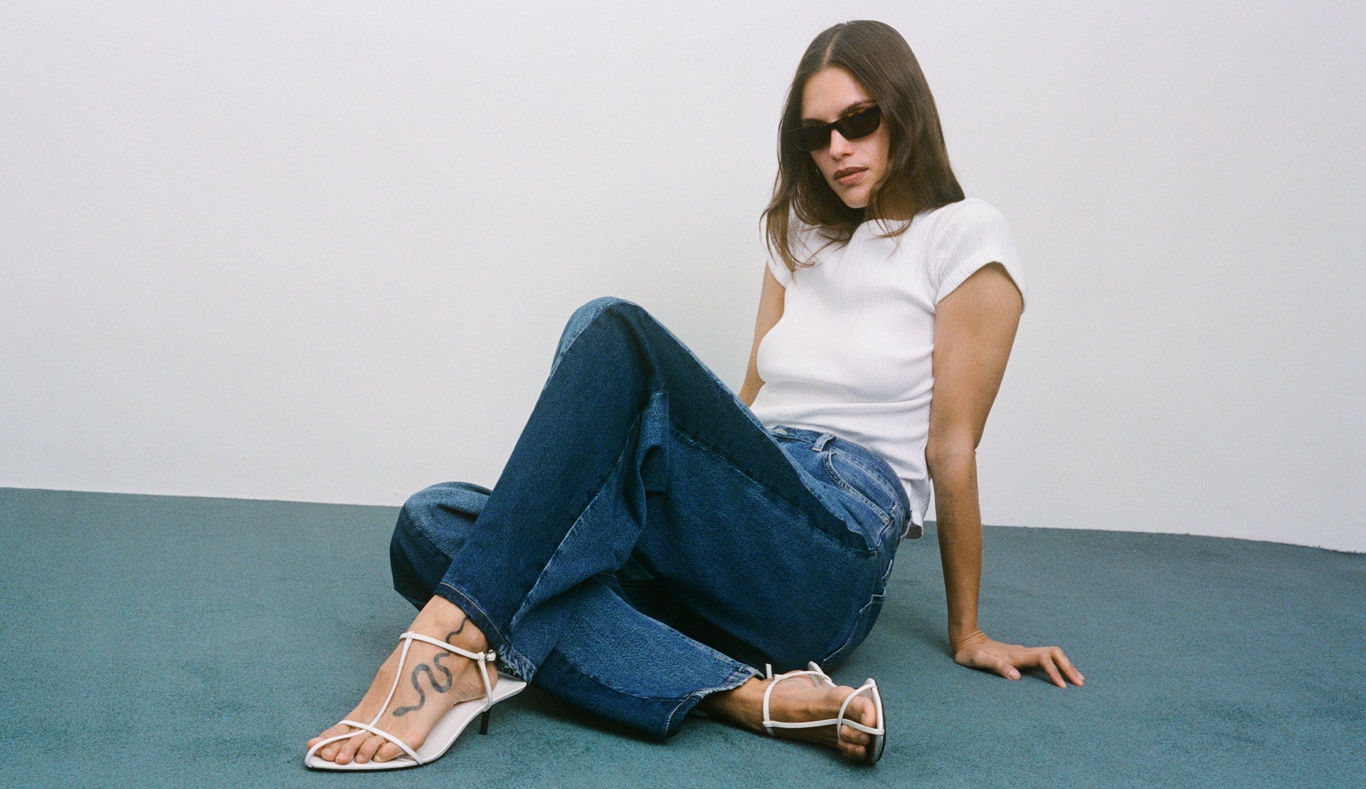 Woman sitting on a blue carpet wearing sunglasses, white shirt, blue jeans, and white heels.
