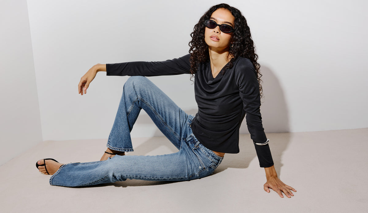 Woman seated in a black cowl-neck long-sleeve top, light-wash flared jeans, black strappy heeled sandals, and oval sunglasses.
