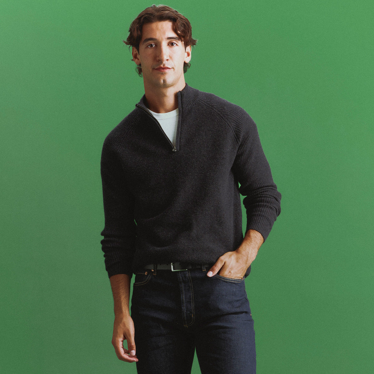 Man in a charcoal ribbed quarter-zip sweater over a white tee and dark jeans, one hand in his pocket, against a green backdrop.