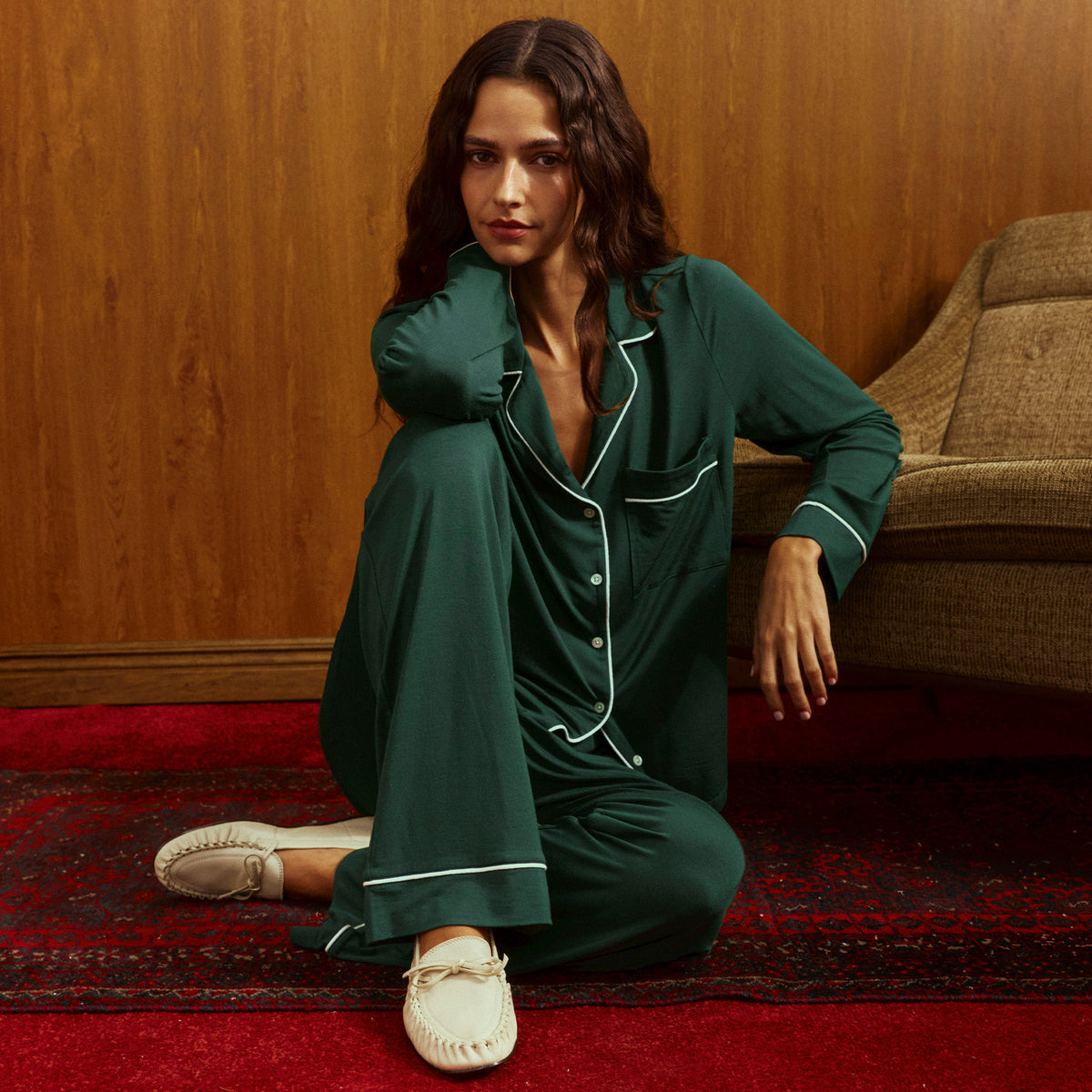 Woman with wavy dark hair sitting on a red patterned rug in green pajamas with white piping and cream bow loafers, leaning against an olive armchair in front of a wood-paneled wall.