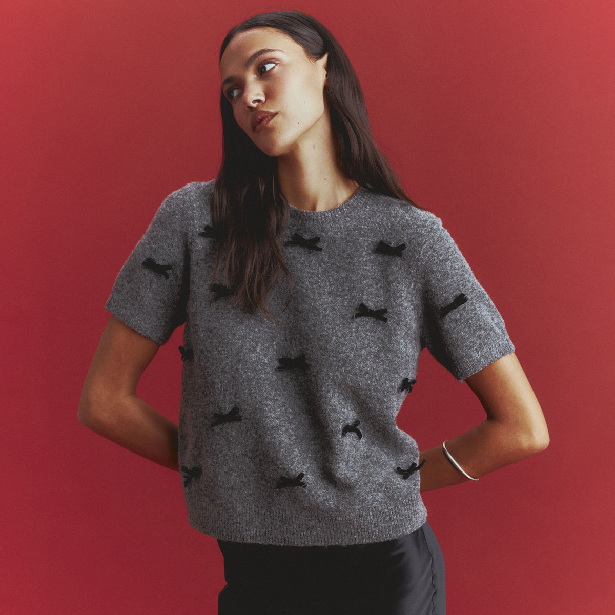 Woman in a gray short-sleeve knit sweater scattered with small black velvet bows, worn with a black skirt and a silver bangle, against a red backdrop.