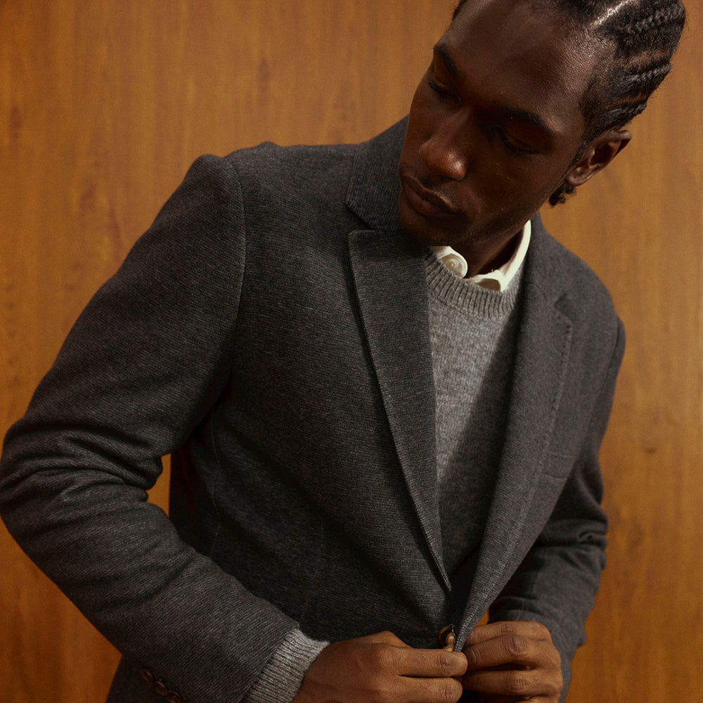 A man wearing a dark gray wool-blend blazer over a gray knit sweater and white collared shirt, standing against a warm wood-paneled background and buttoning his blazer.
