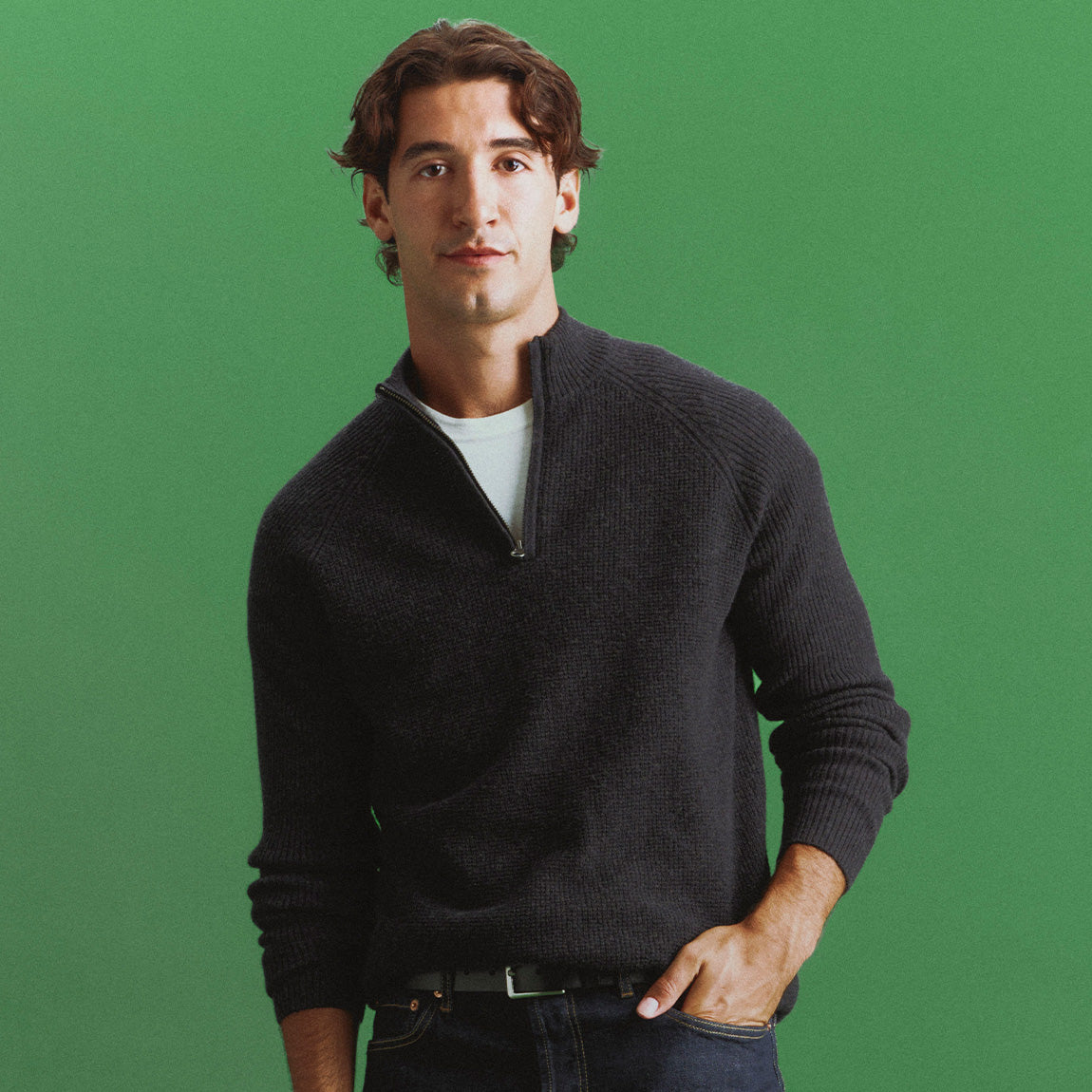 Man in a charcoal ribbed quarter-zip sweater over a white tee and dark jeans, one hand in his pocket, against a green backdrop.