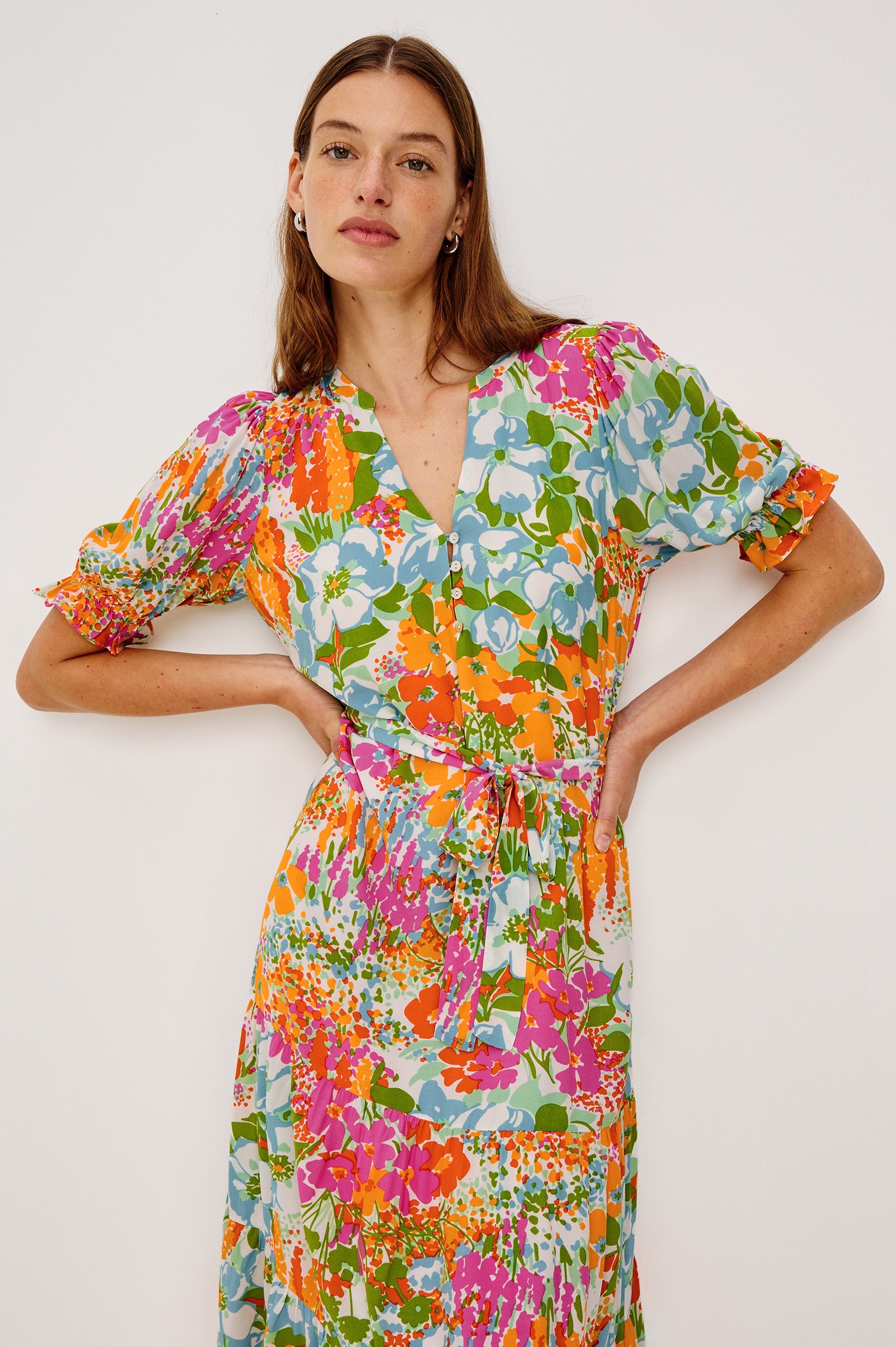 Eliana Midi Dress with Elbow-Length Sleeves with Shirred Cuffs, a V Neck, Buttons Down the Bodice, a Fabric Tie at the Waist  and a Tiered Skirt in Day Garden Colorway, a Floral Pattern in Blue, Pink and Oranges on White - Close up View Featured on Model