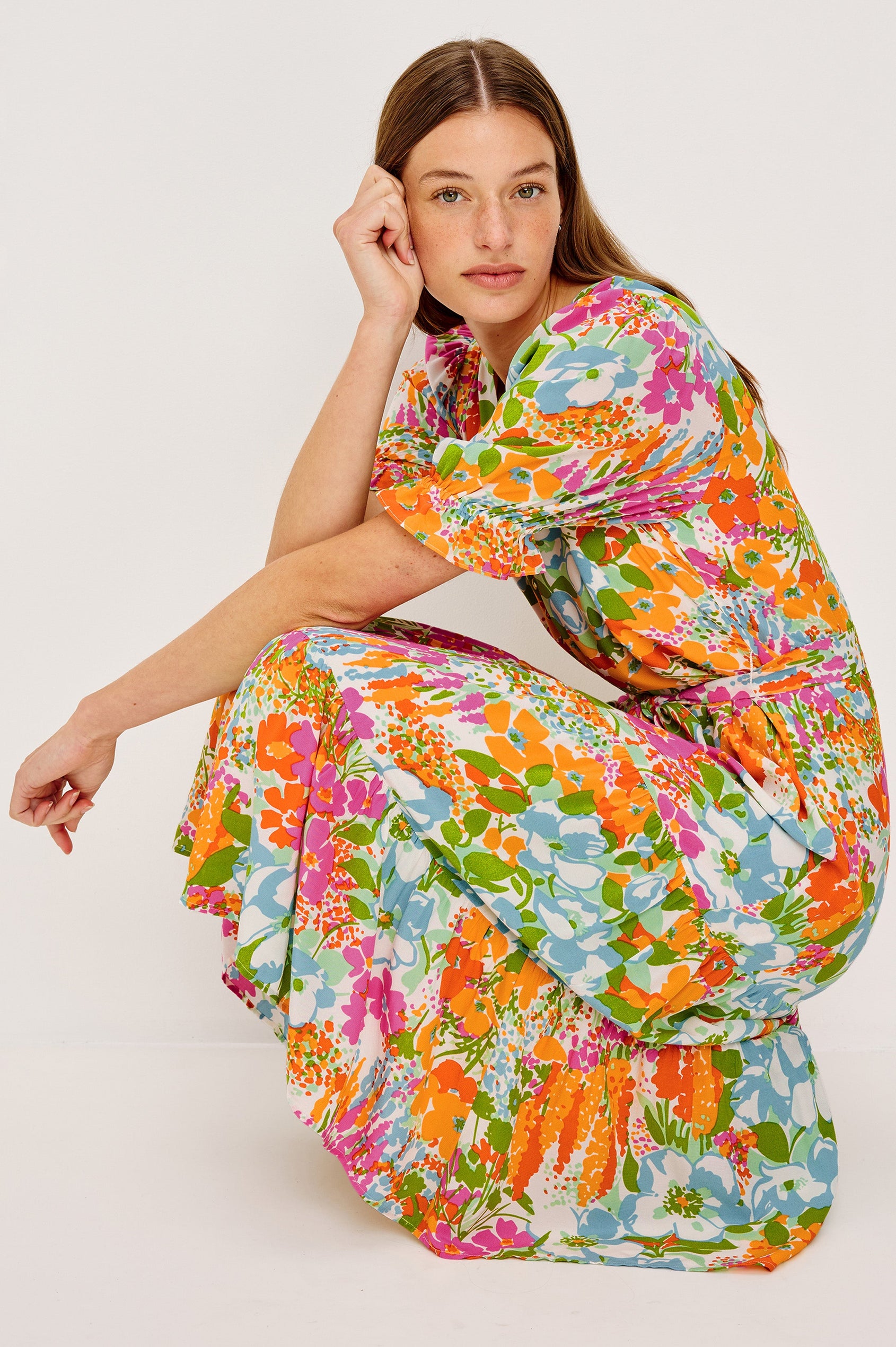 Eliana Midi Dress with Elbow-Length Sleeves with Shirred Cuffs, a V Neck, Buttons Down the Bodice, a Fabric Tie at the Waist  and a Tiered Skirt in Day Garden Colorway, a Floral Pattern in Blue, Pink and Oranges on White - Front View Featured on Model in Squatting position