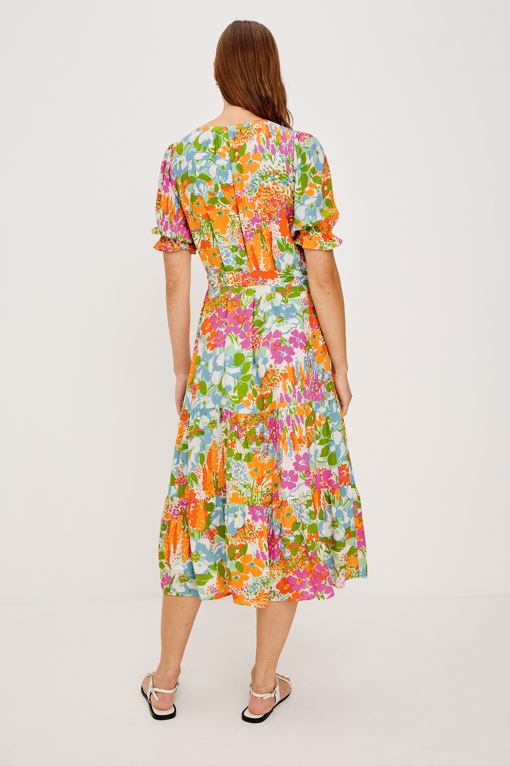 Eliana Midi Dress with Elbow-Length Sleeves with Shirred Cuffs, a V Neck, Buttons Down the Bodice, a Fabric Tie at the Waist  and a Tiered Skirt in Day Garden Colorway, a Floral Pattern in Blue, Pink and Oranges on White - Back View Featured on Model
