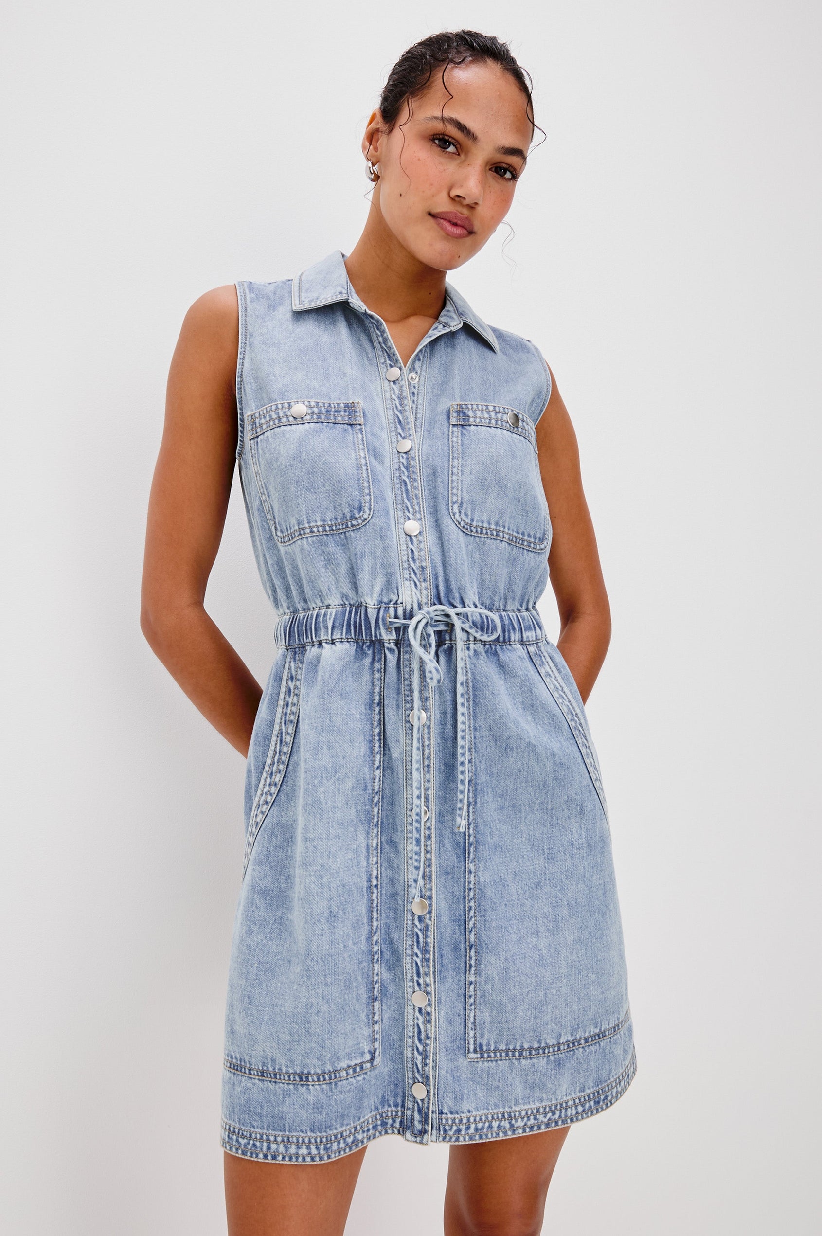 Erica Lightweight Denim Mini Dress with a Exposed Button Front, Encased Drawstring Waistband, Front Patch Pockets, Chest Pockets, Sleeveless and Collared in Faded Indigo Colorway. Front View Shown on Model.