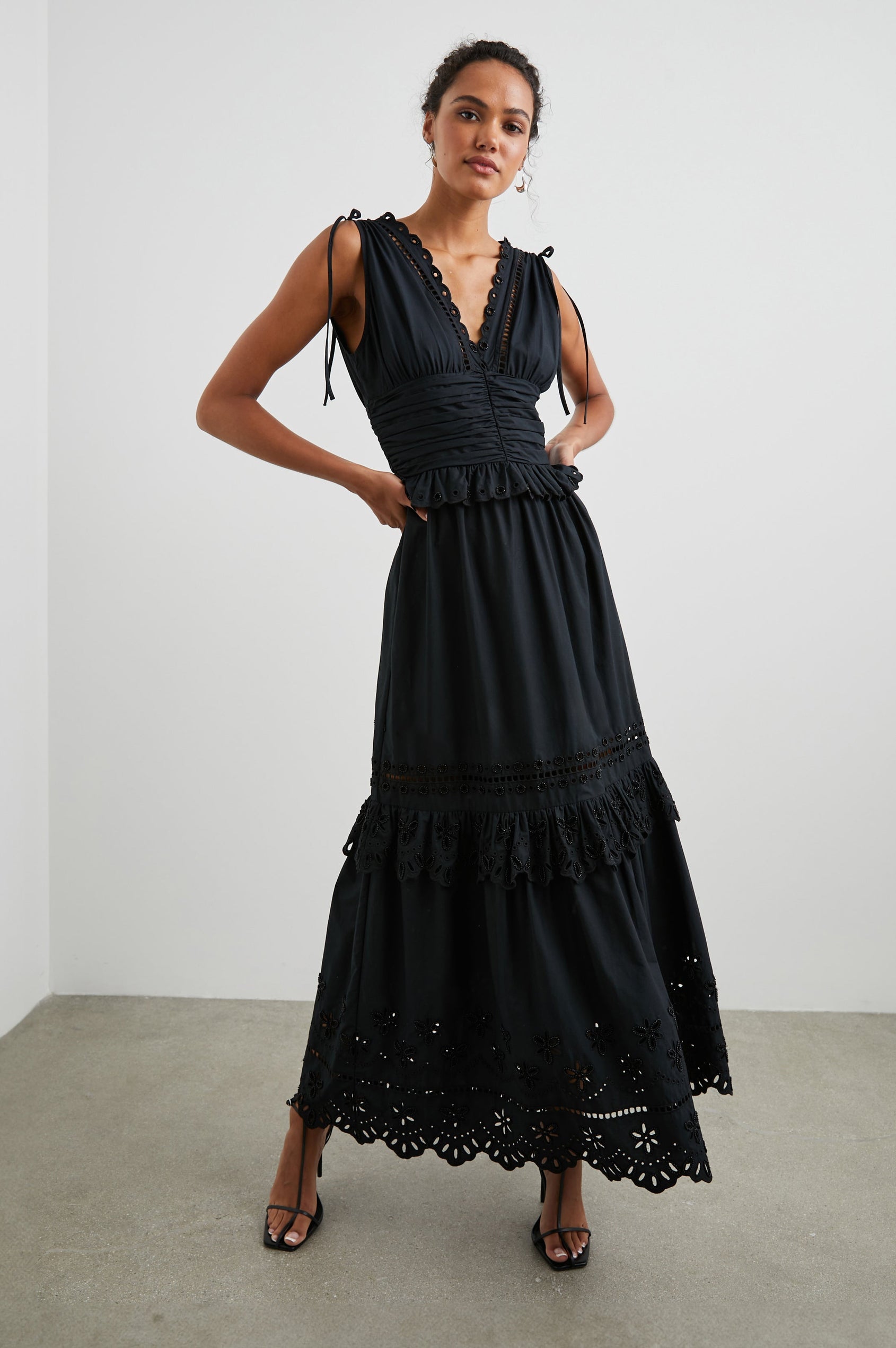 Esmeralda Sleeveless Maxi Dress with a Deep V Neck, a Ruched Waistband, Drawstrings Across the Shoulder Straps, Scalloped Trim with Cutouts on the Bodice, a Tiered Skirt, and an Eyelet Design along the Hem in Black Colorway - Front Full Body View Featured on Model 