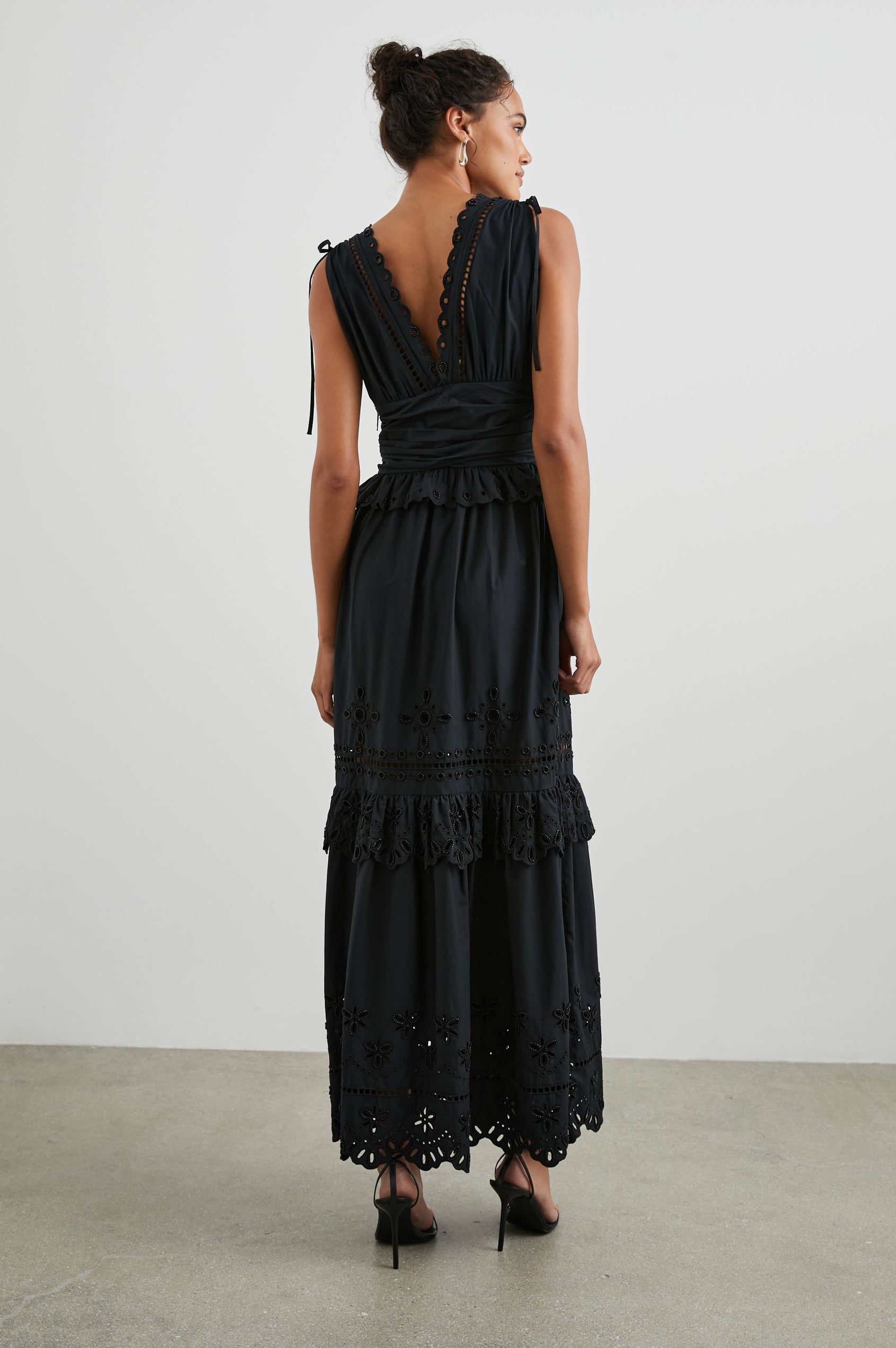 Esmeralda Sleeveless Maxi Dress with a Deep V Neck, a Ruched Waistband, Drawstrings Across the Shoulder Straps, Scalloped Trim with Cutouts on the Bodice, a Tiered Skirt, and an Eyelet Design along the Hem in Black Colorway - Back Full Body View Featured on Model 