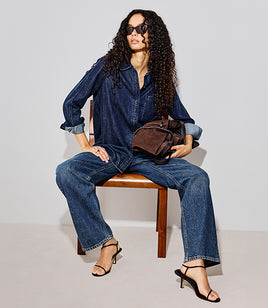 Woman with dark curly hair wearing an all denim outfit with black heels and a brown suede bag