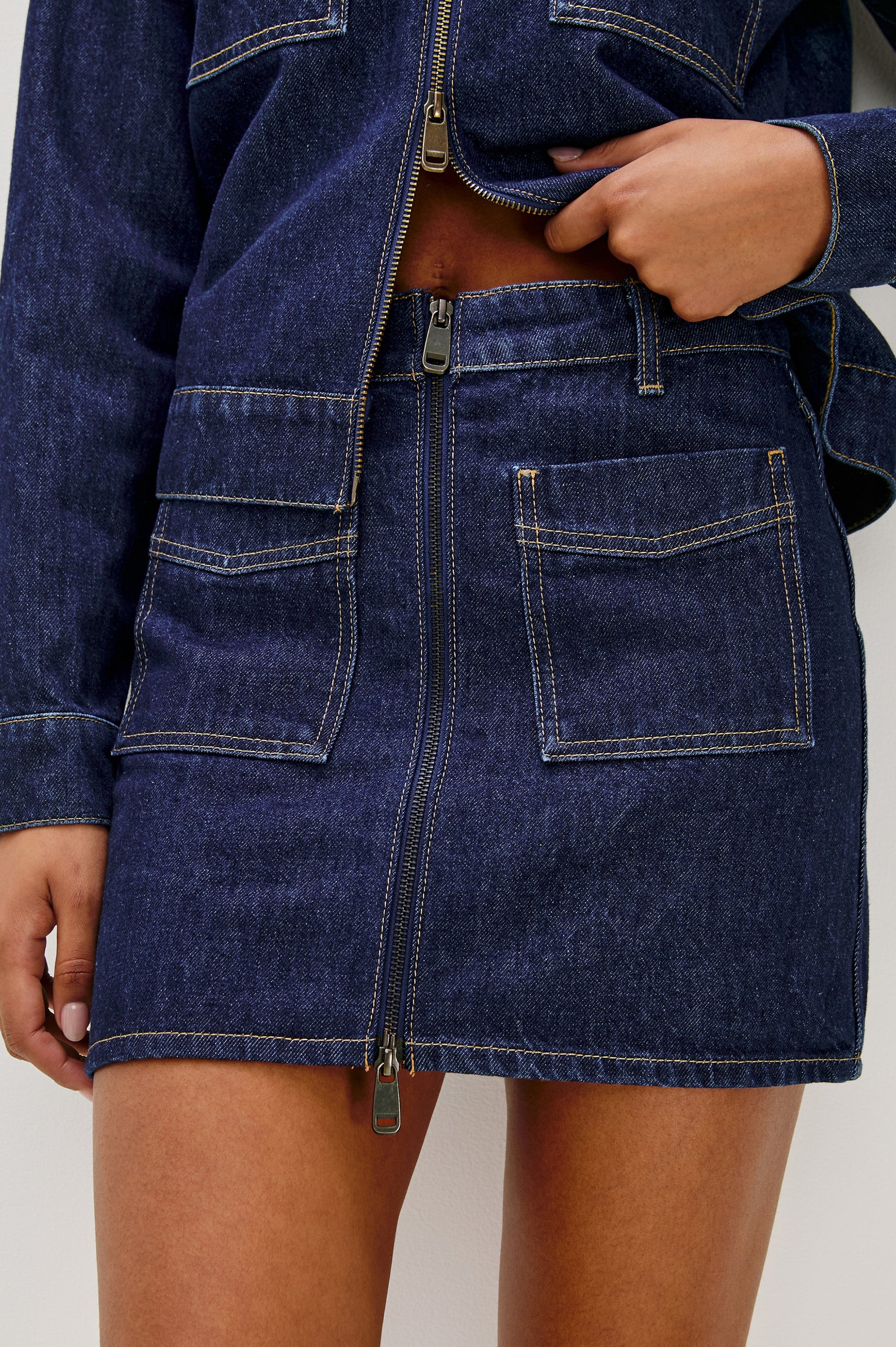 The Fallbrook is a mini zip front denim skirt with beltloops, front patch pockets and back pockets in Deep Indigo colorway. Front detail view featured on model.