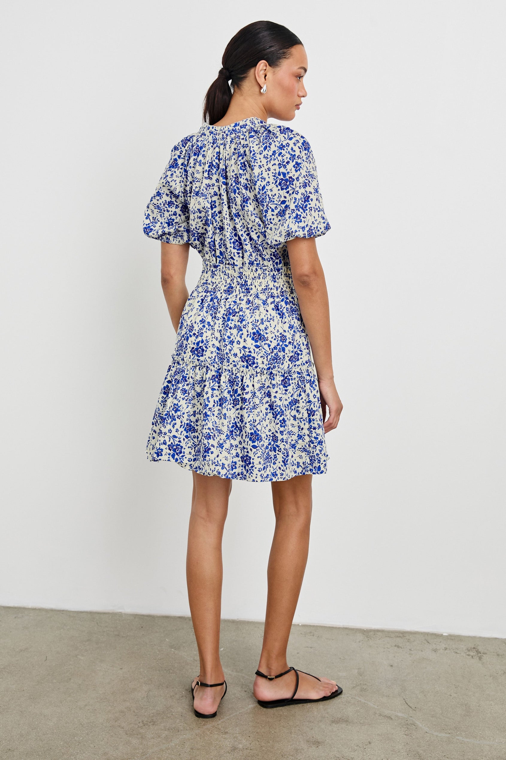 Fiorella Above-Knee Dress with Short Elasticated Puff Sleeves, Split V-Neck with Self Ties, and an Elasticated Ruched Waist Detail in Blue Marguerite Colorway. Full Body Back View Shown on Model.