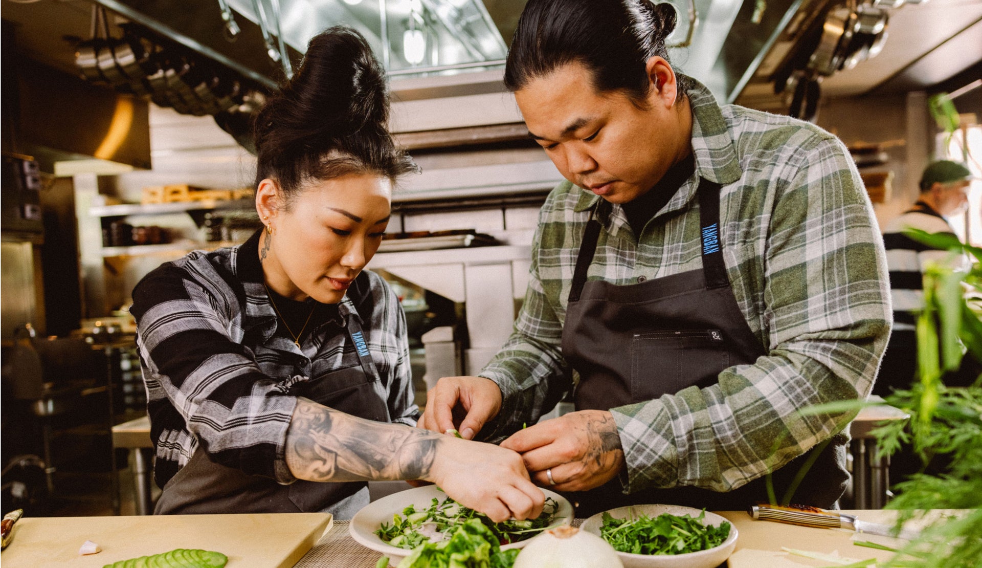 MALE AND FEMALE COOKING. FEMALE IS WEARING HUNTER SHIRT (LONG SLEEVE BUTTON UP SHIRT) IN COLOR BLACK CINDER (BLACK AND GREY PLAID). MALE IS WEARING LENNOX SHIRT (LONG SLEEVE BUTTON DOWN) IN COLOR GREEN HOLLOW GREY MELANGE (GREEN AND GREY PLAID)