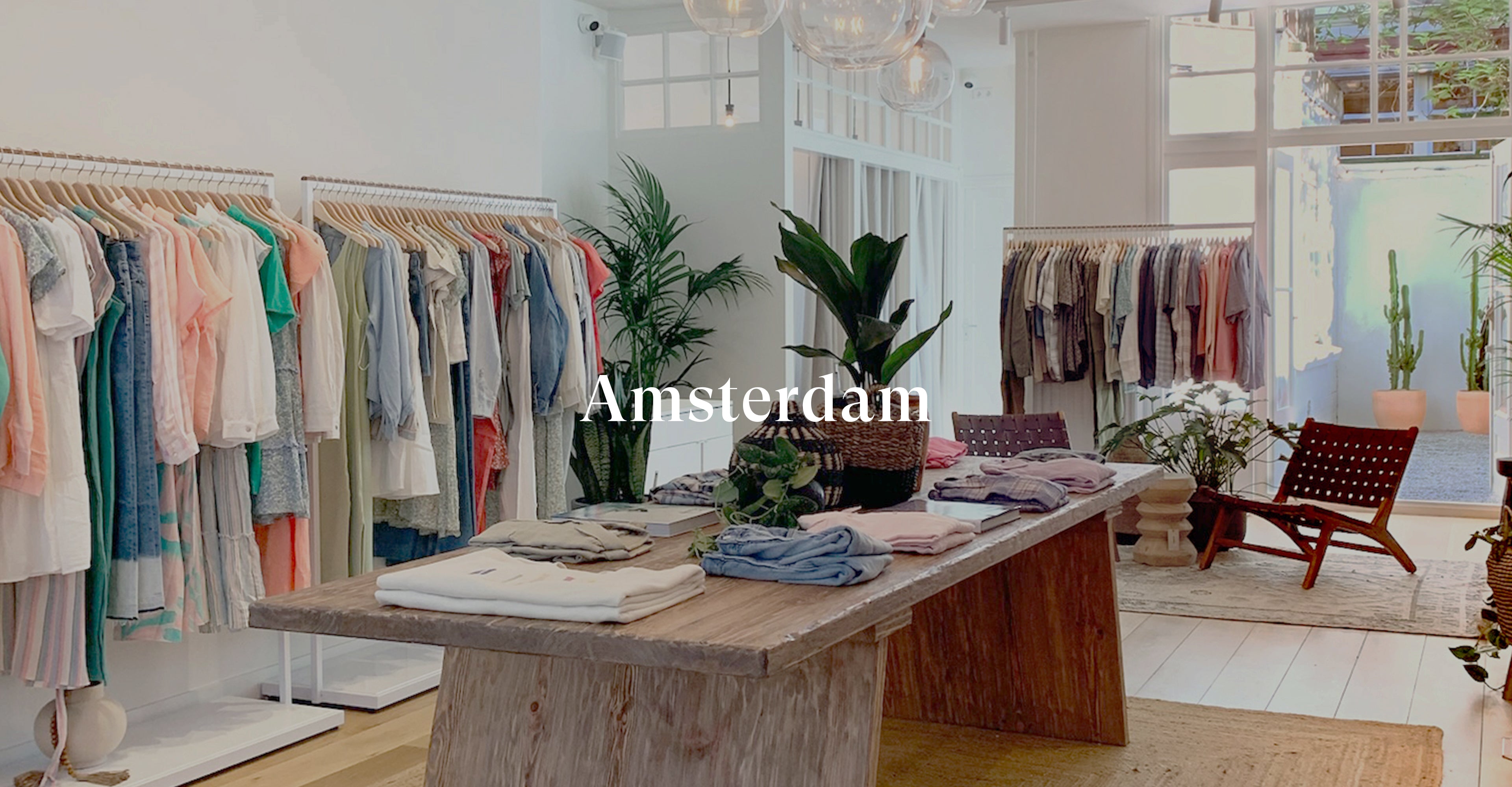 INSIDE OF AMSTERDAM STORE SHOWING LARGE TABLE WITH CLOTHING ON TOP OF IT IN MIDDLE OF ROOM AND CLOTHING RACKS FILLED WITH CLOTHING AGAINST EACH WALL