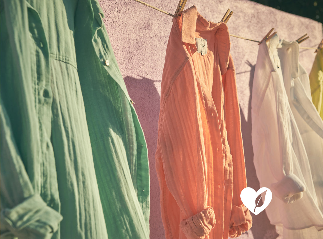 THREE ELLIS SHIRTS (COTTON LONG SLEEVE BUTTON UP IN A LOOSE FIT WITH COLLAR) HANGING ON A CLOTHES LINE. EACH SHIRT IS A DIFFERENT COLOR (FROM LEFT TO RIGHT COLORS ARE GREEN, ORANGE, WHITE)