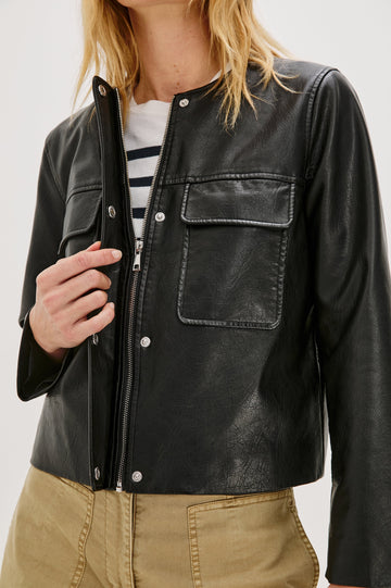 Inka, a cropped, boxy faux leather jacket in a black, slightly distressed finish. The design includes classic chest pockets and a structured silhouette. Close Up View featured on model, Zipped