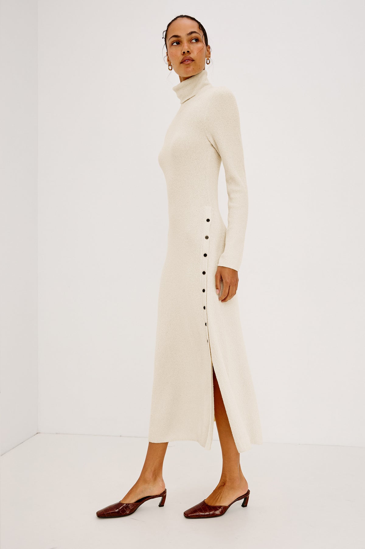 Kat Maxi Dress in Ribbed Cotton Blend With Foldover Turtleneck Button Neckline, Long Sleeves, Fitted Silhouette, Long Sleeves, Side Placket, and Subtle Flare at Hem in Ivory Colorway - Full Body Side View Featured on Model