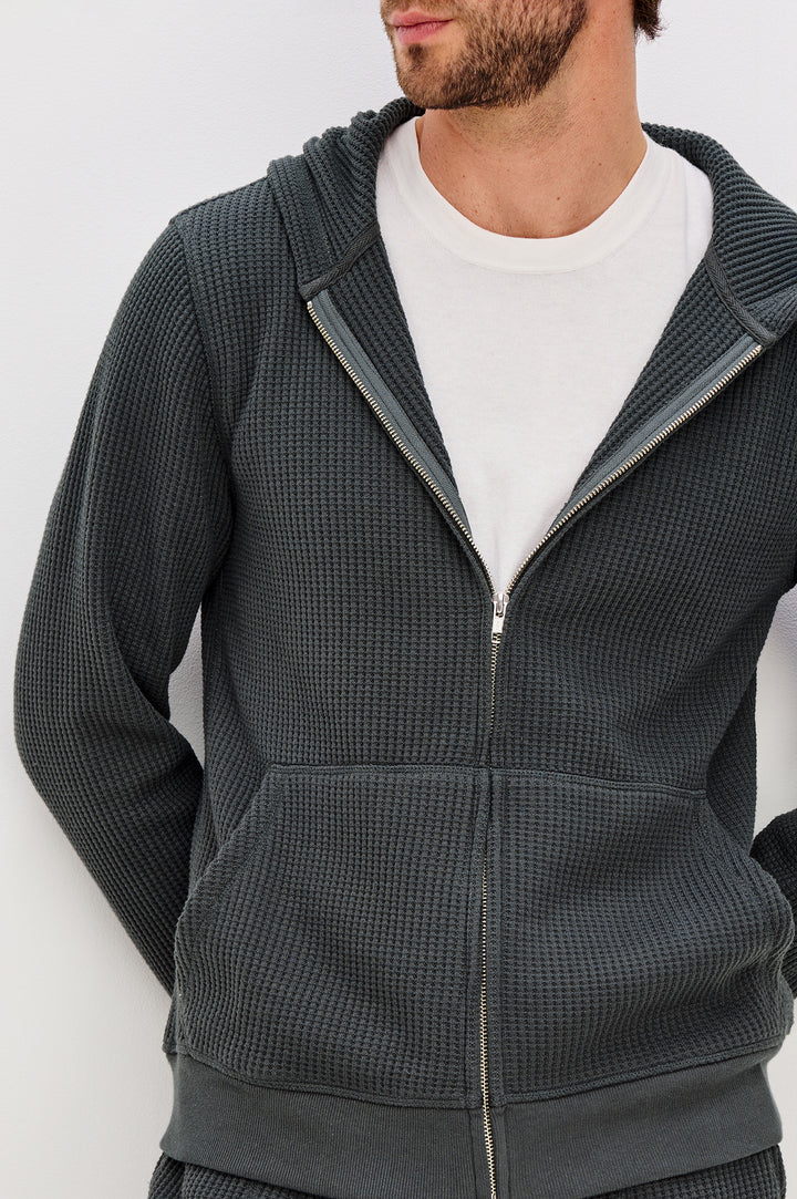 Kenton is a zip front hooded sweatshirt with front patch pockets and ribbed cuffs in Ink colorway. Front detail view featured on model.