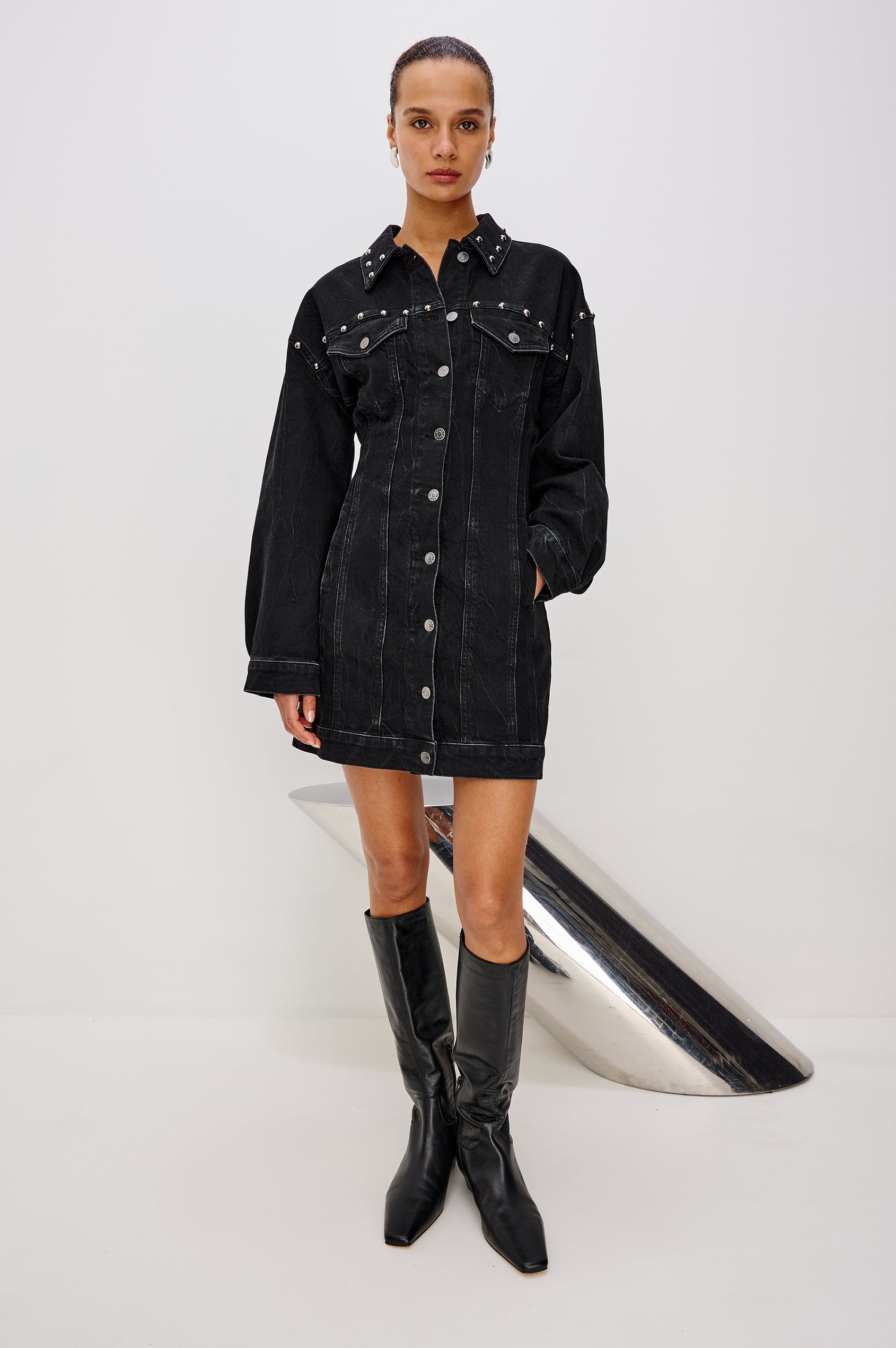 Ladera is a trucker style denim mini dress with long sleeves, drop shoulders, relaxed balloon sleeves, nail-back studs, and front button closures, with a subtle A-line shape and adjustable back strap in Black Stone Studded colorway. Front full body view featured on model.