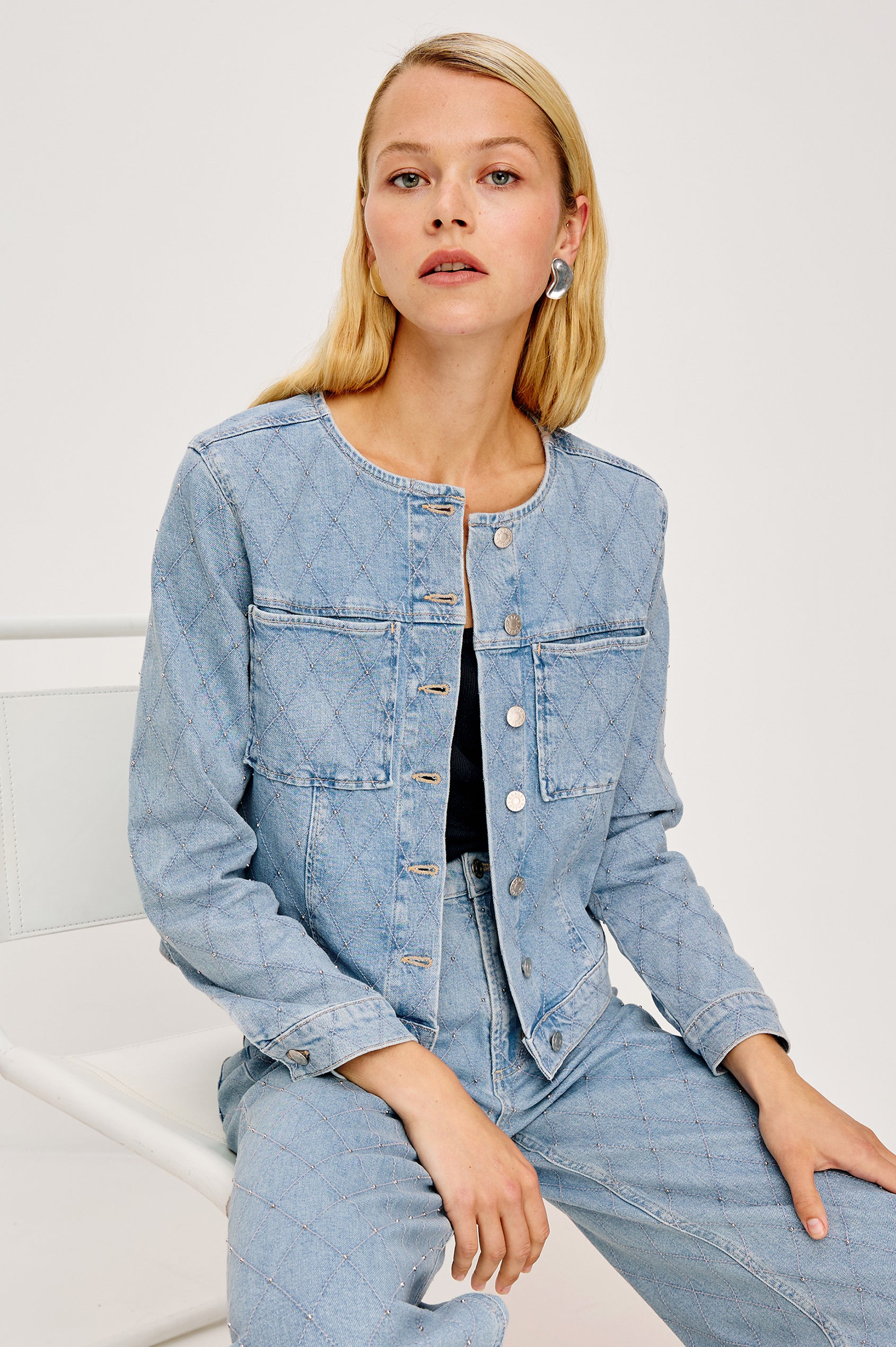 Lido Lady Jacket in Light Stretch Denim With Cropped Boxy Fit, Collarless Neckline, Front Buttons, Long Sleeves, and Chest Patch Pockets in Quilted Stud Colorway - Full Body Front View Featured on Model Sitting