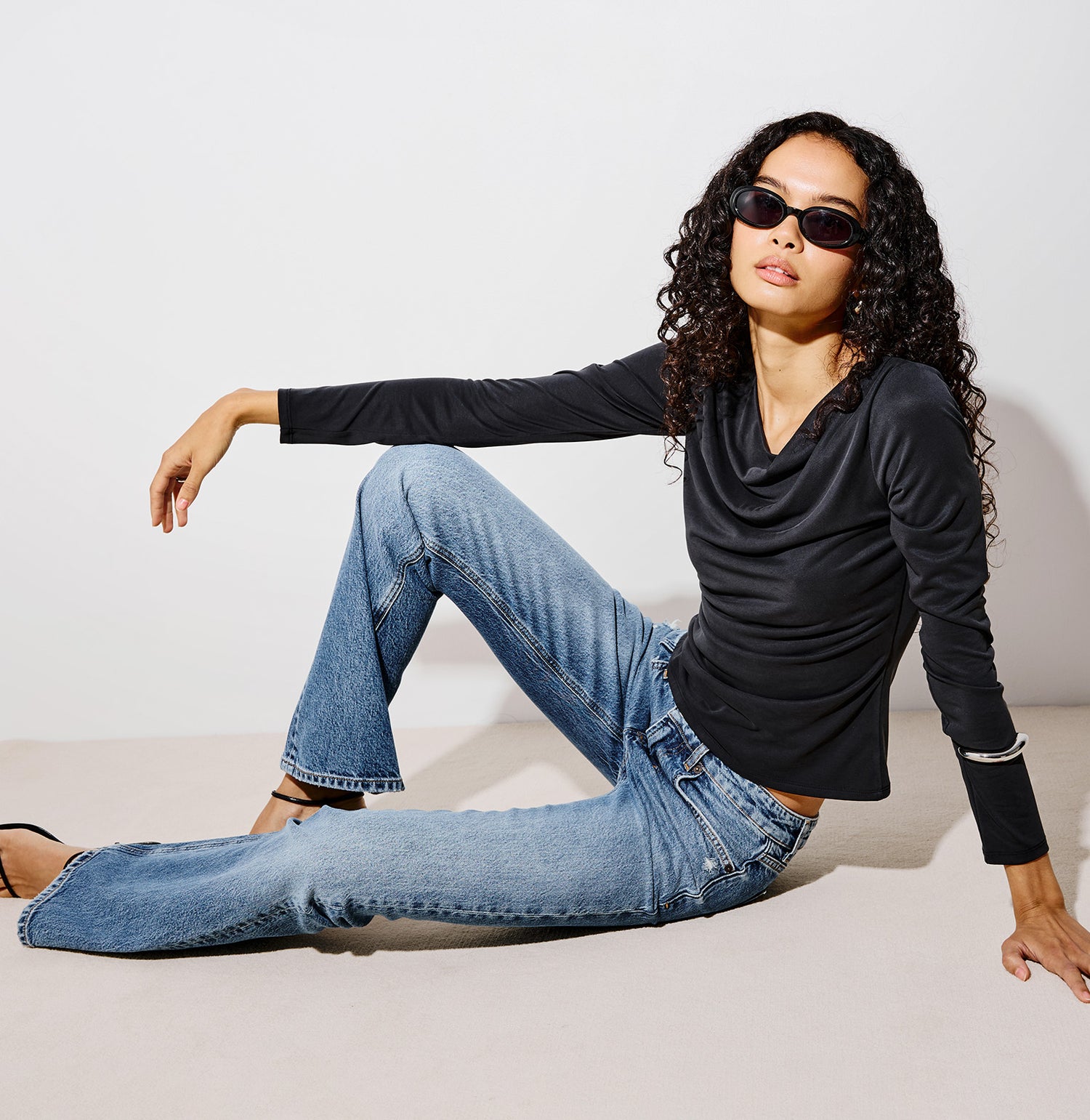 A woman wearing a black long-sleeve cowl-neck top, blue jeans, and black sunglasses sits on a light beige floor against a white wall.