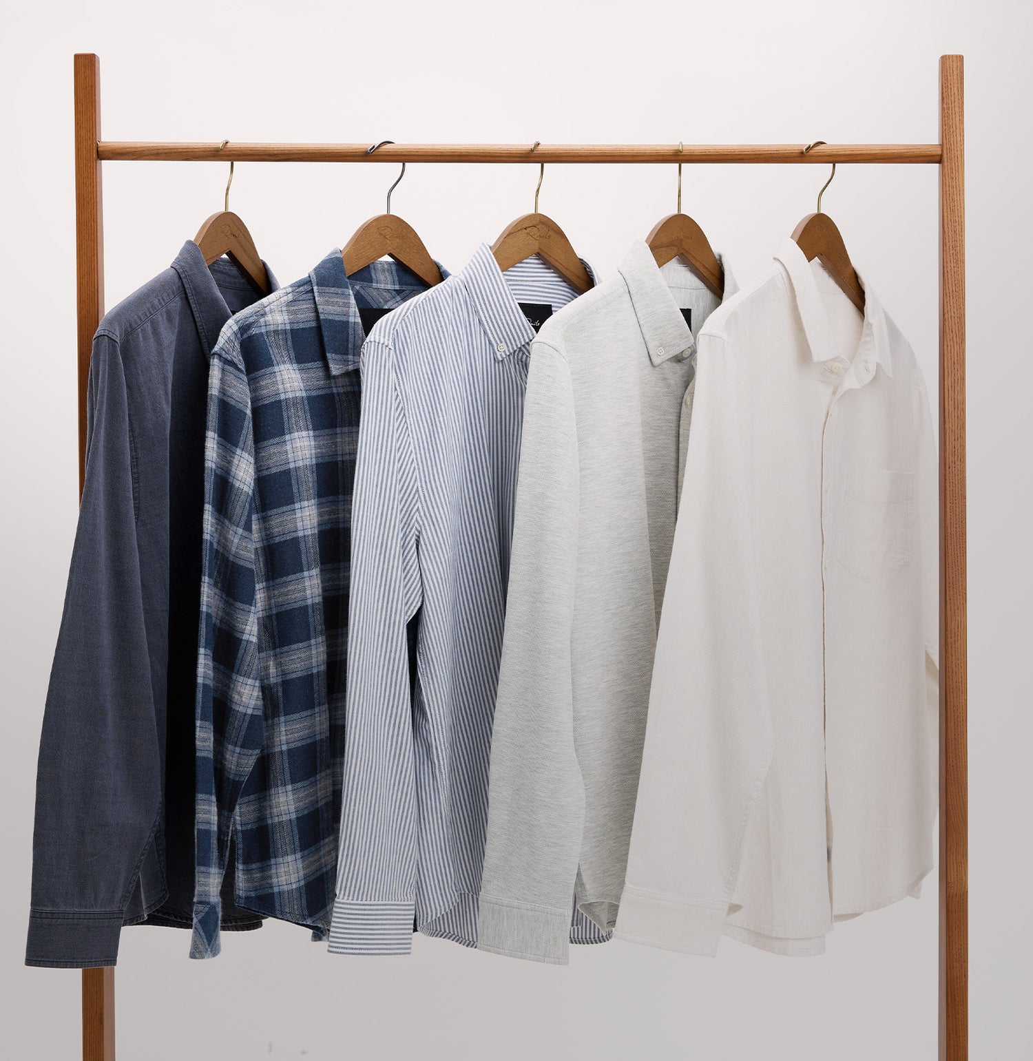 Five long-sleeve button-up shirts in different colors and patterns hang on a wooden clothing rack against a plain white wall. The shirts include dark navy, blue plaid, light blue, light gray, and off-white.