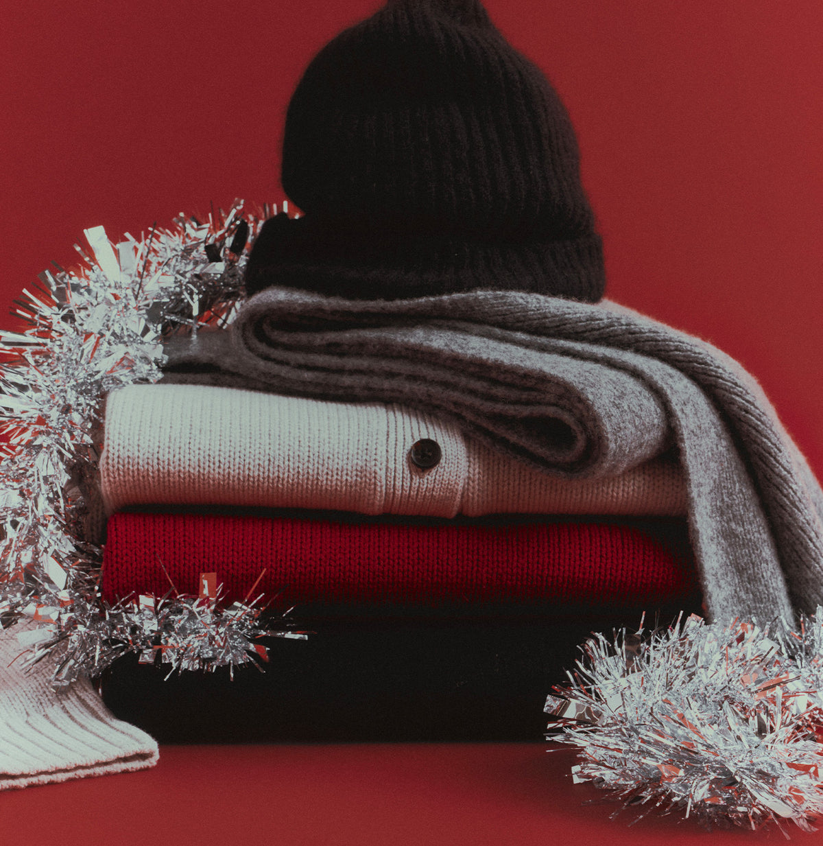 A stack of folded knit garments in gray, cream, and red, styled with silver tinsel against a solid red background.