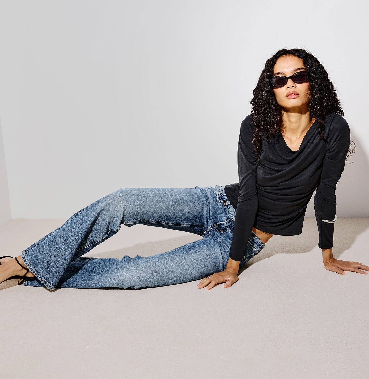 A model wearing a black long-sleeve cowl-neck top, light-wash blue flare jeans, and black sandals, posing on a minimalist light gray studio floor and wall.