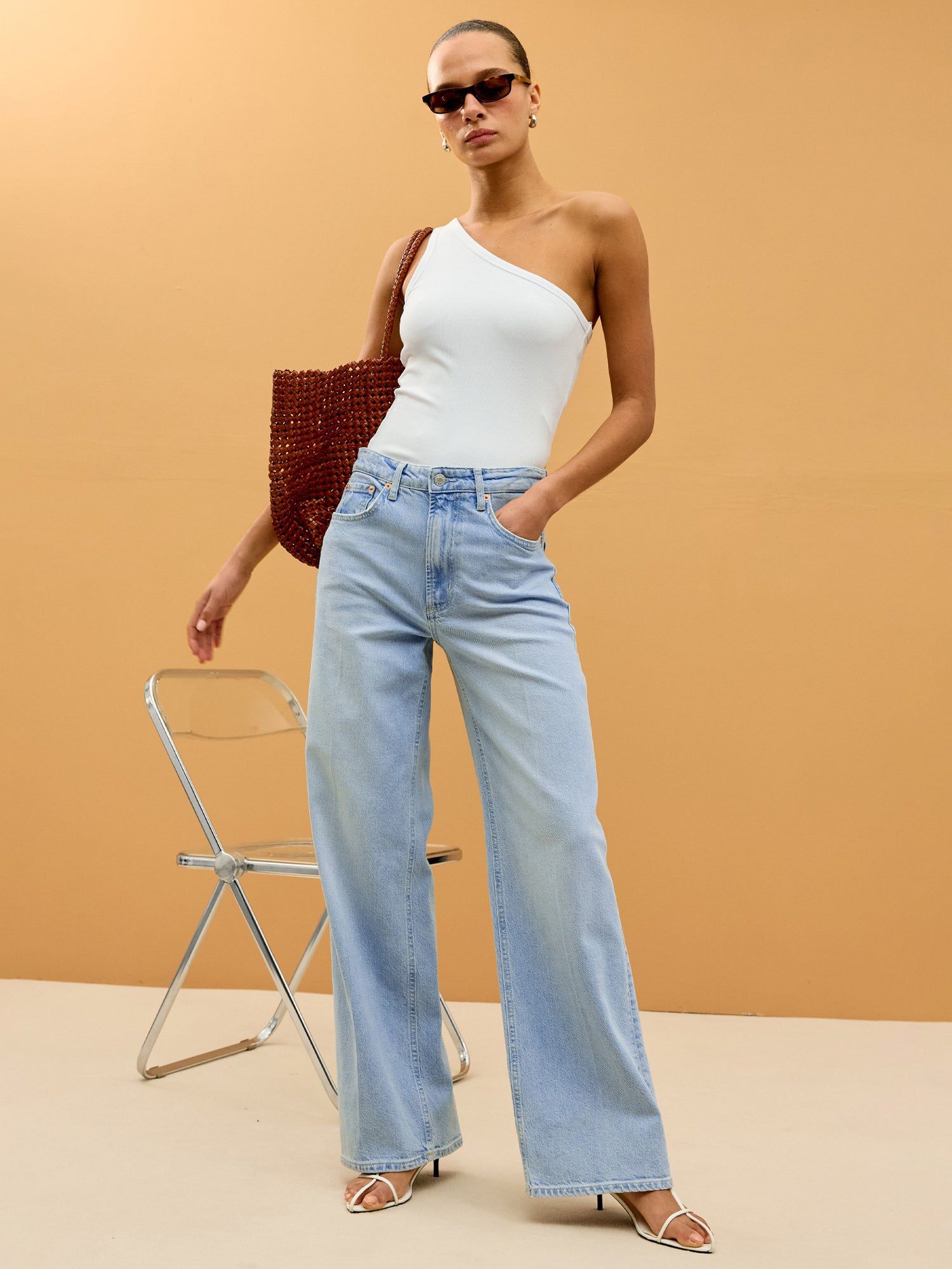 Woman wearing a white one-shoulder top and light blue jeans against a beige background