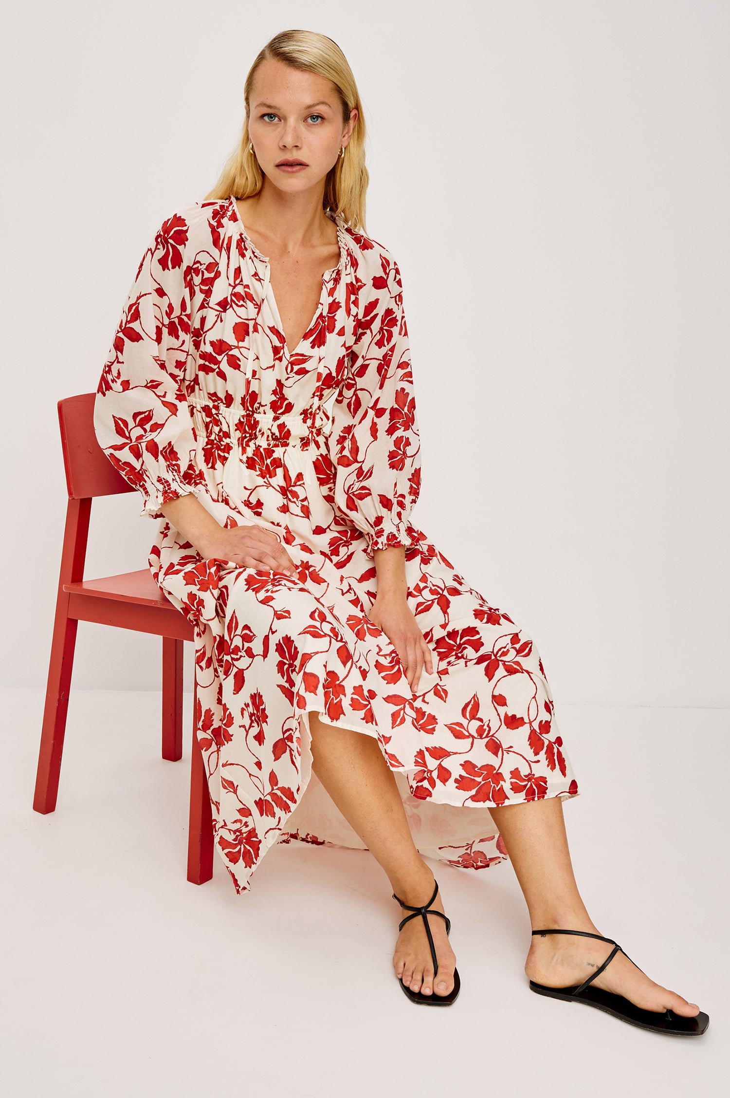 Matia Maxi Dress in Cotton With Banded Ruffle V-Neckline With Self-Ties Elastic Waist and Long Raglan Puff Sleeves in Red Jasmine Colorway - Full Body Front View Featured on Model Sitting