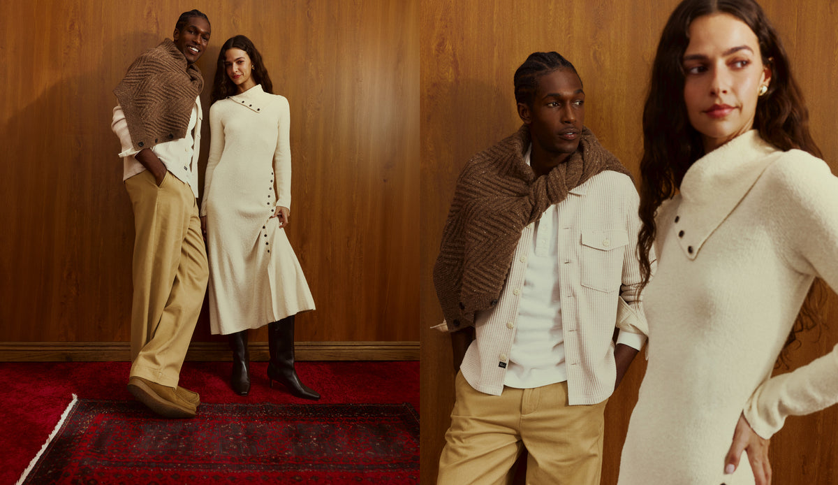 A man and a woman pose together in a warm, rustic indoor setting with a wood-paneled wall and a red patterned rug. The man wears a brown textured knit shawl over a white shirt and tan pleated trousers.