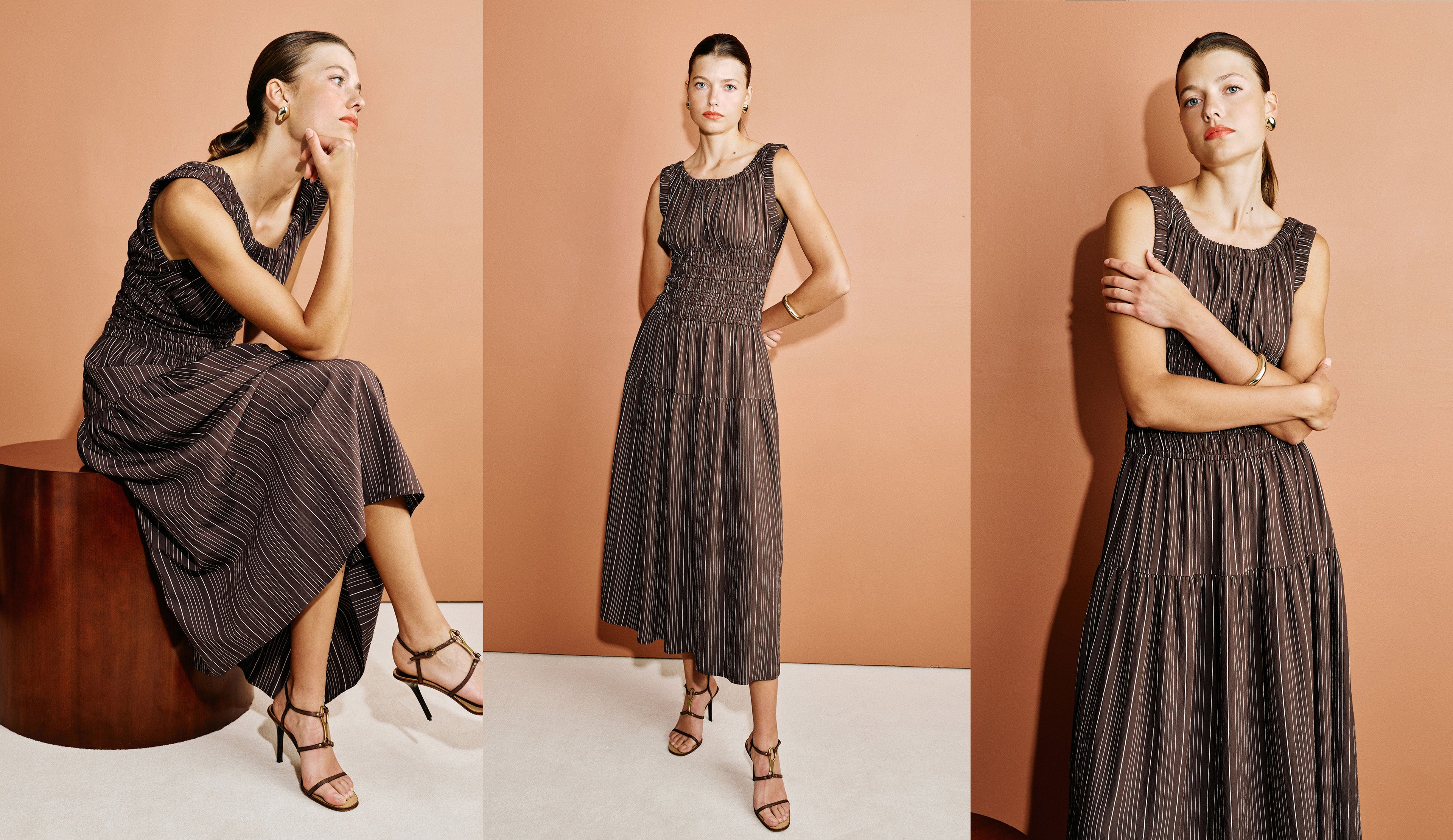 A woman modeling a sleeveless pleated midi dress in a dark brownish-gray color, shown in three poses against a warm peach studio backdrop: seated on a wooden stool, standing with one hand on her hip, and standing with arms crossed.