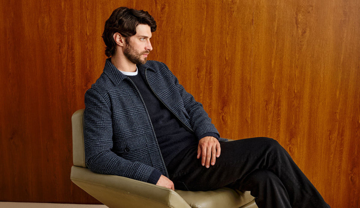 A man with dark hair and a beard sits in a beige armchair against a warm wood-paneled wall, wearing a dark blue textured jacket over a black shirt and black trousers.